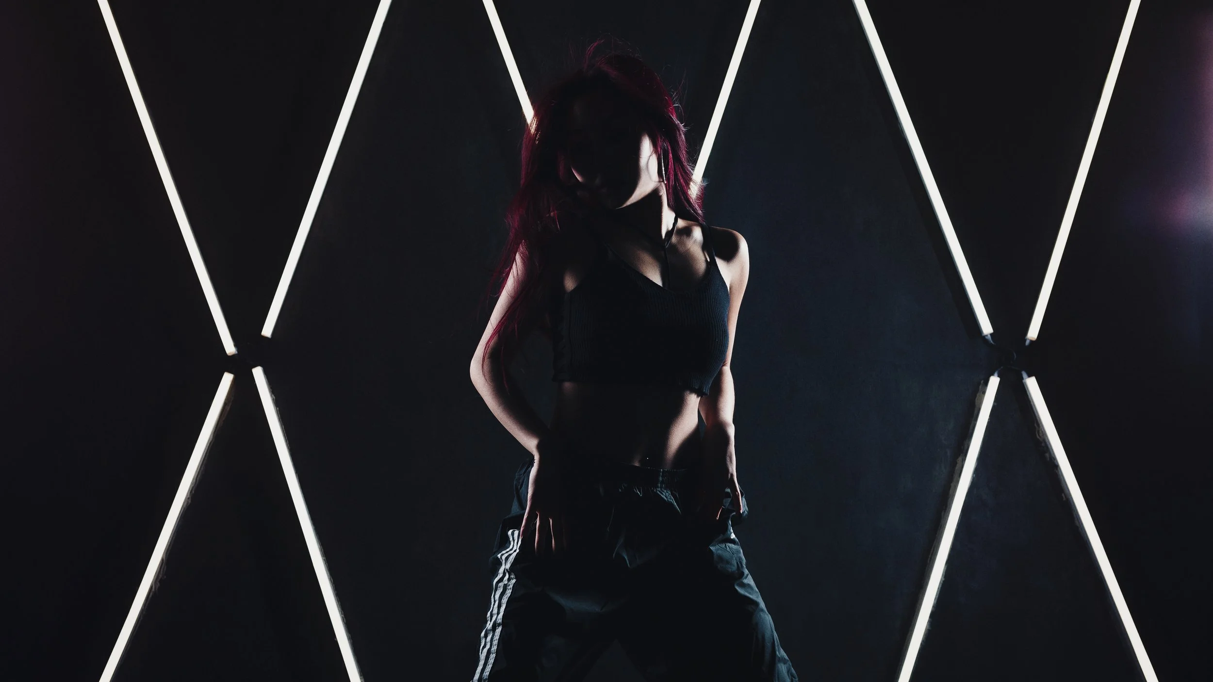 A woman with red hair posing in front of a dark background with white neon light lines forming geometric patterns, wearing a black crop top and black pants.