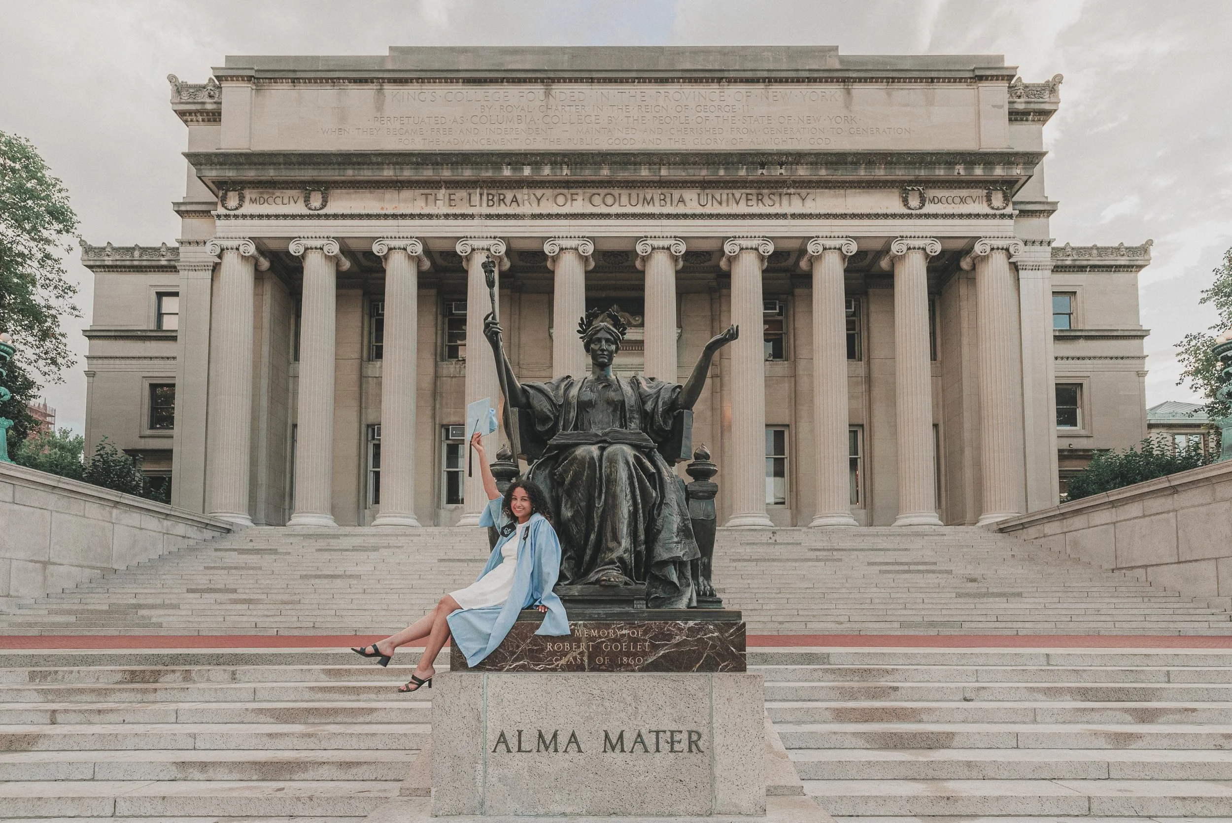 A woman in a graduation gown sitting on the steps of Columbia University's library, holding a blue cap in front of the university's historic building with columns and statues.