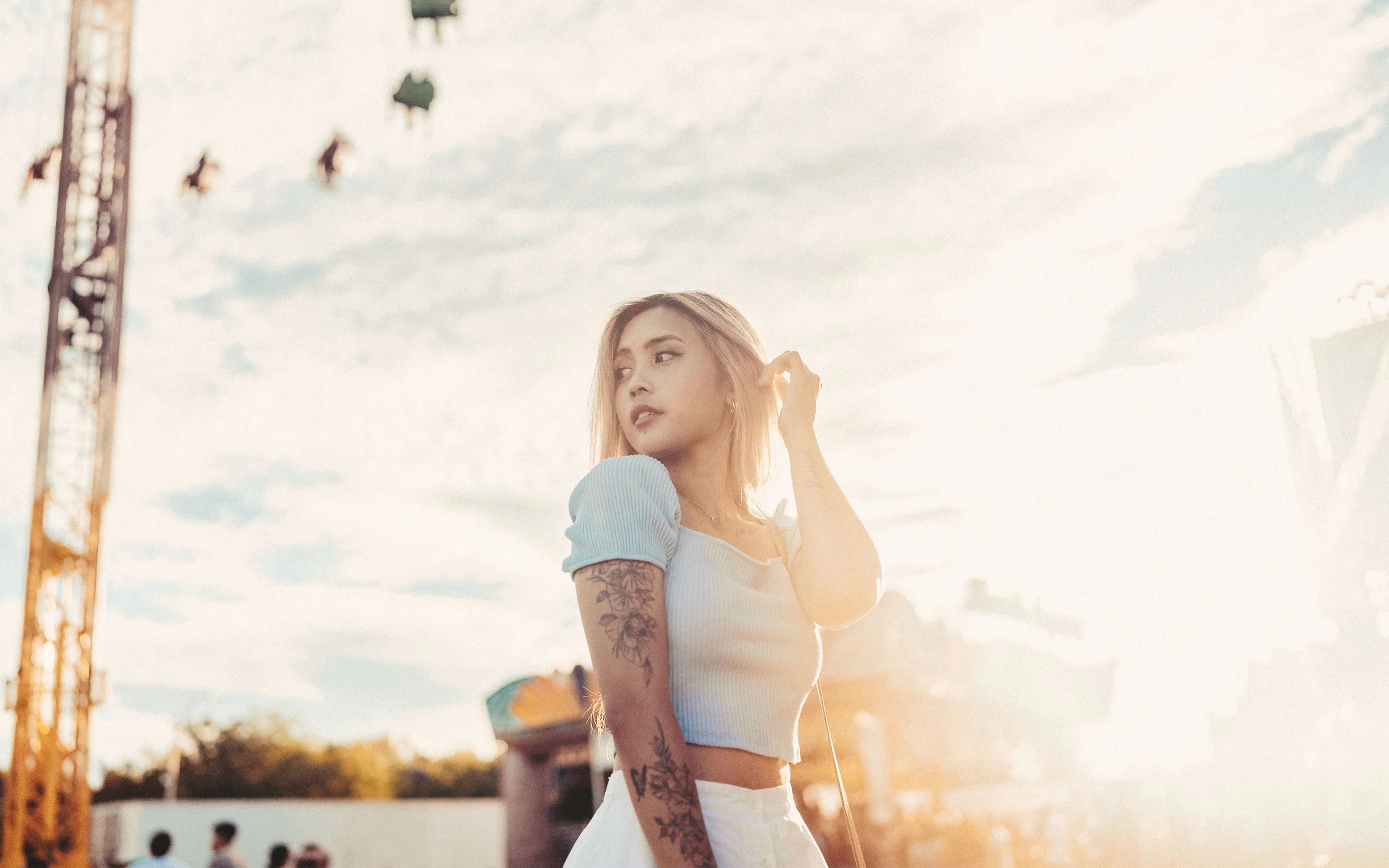 A young woman with blonde hair and tattoos on her arm, wearing a light-colored crop top, standing outdoors during the daytime with a cloudy sky in the background.