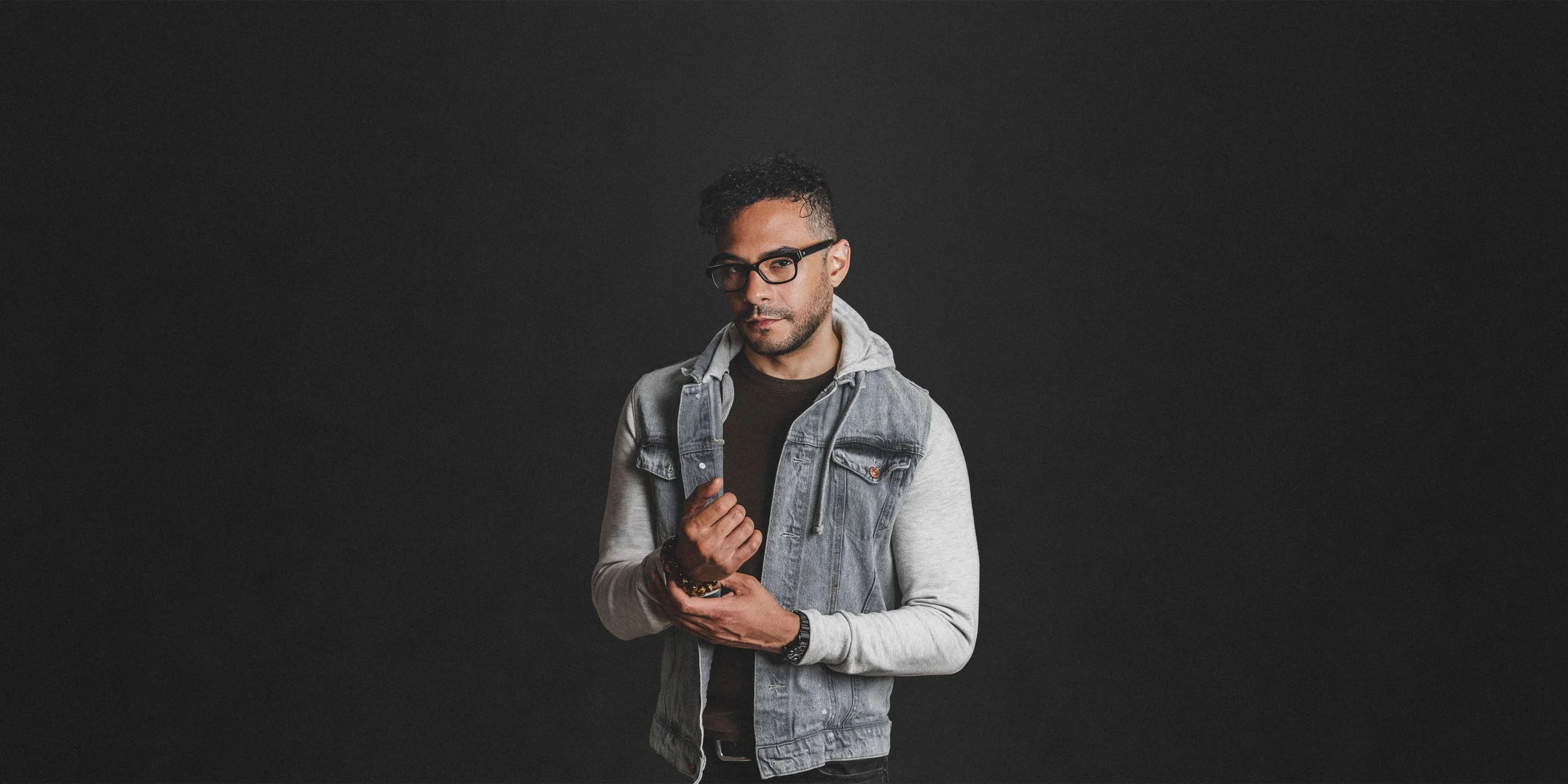 A young man with glasses and curly hair, wearing a gray hoodie under a denim vest, stands against a black background. He is looking at the camera with a slight smirk, holding his left hand with a beaded bracelet visible.