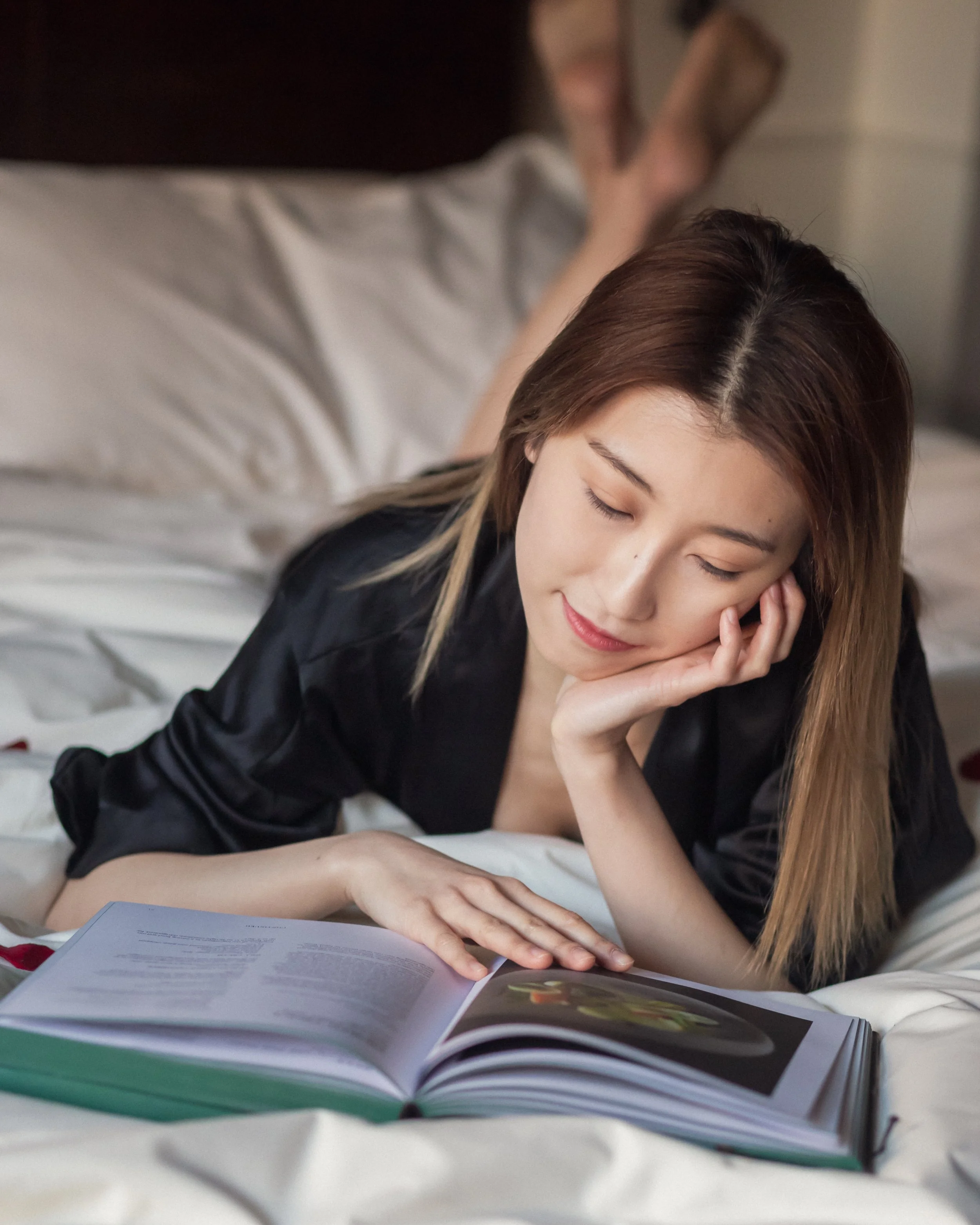 A young woman with long brown hair sitting on a bed, reading a magazine with her eyes closed and resting her face on her hand.