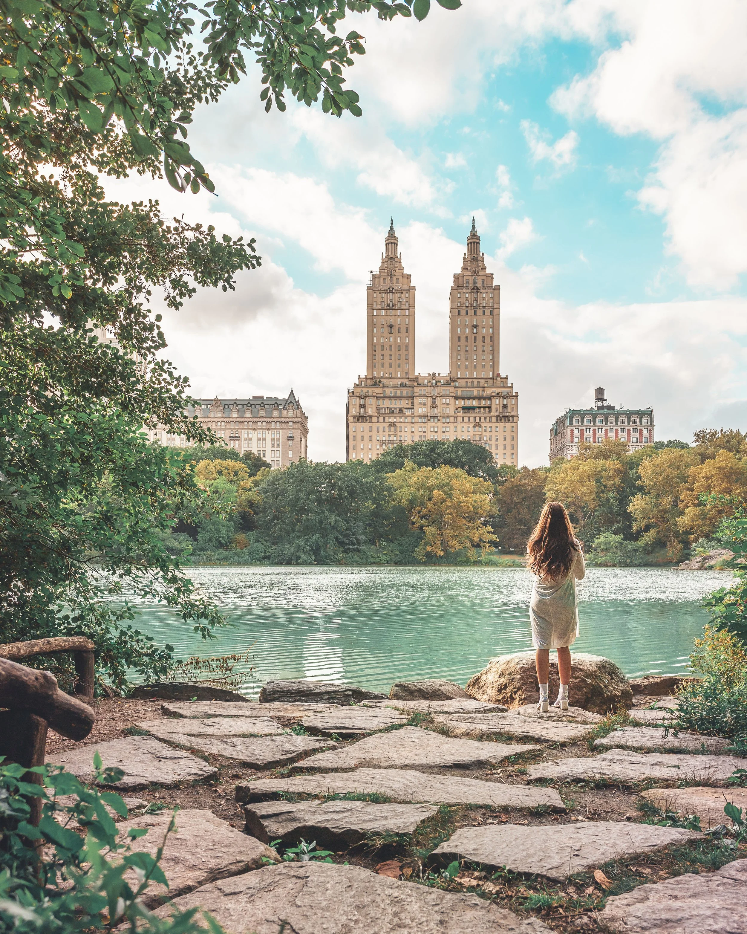 A woman standing on rocks by a lake with trees and buildings, including the iconic twin-towered skyscraper, in the background on a partly cloudy day.
