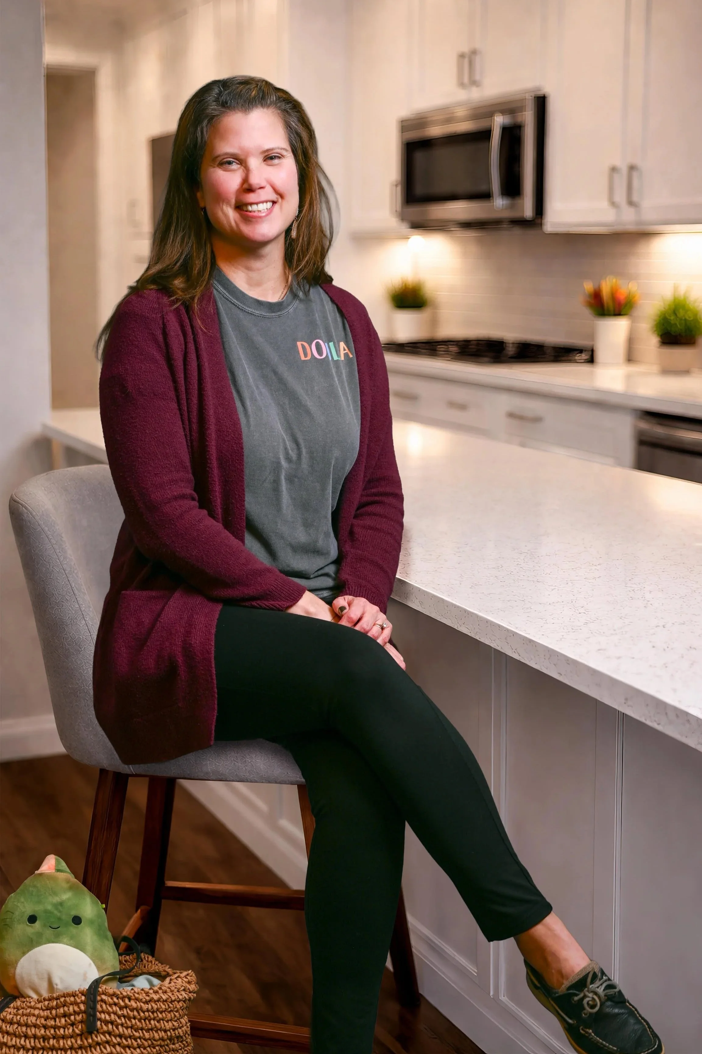A woman with brown hair sitting on a kitchen stool, smiling, wearing a gray 'DOLIA' t-shirt and a maroon cardigan, in a modern kitchen with white cabinets, a microwave, and potted plants.