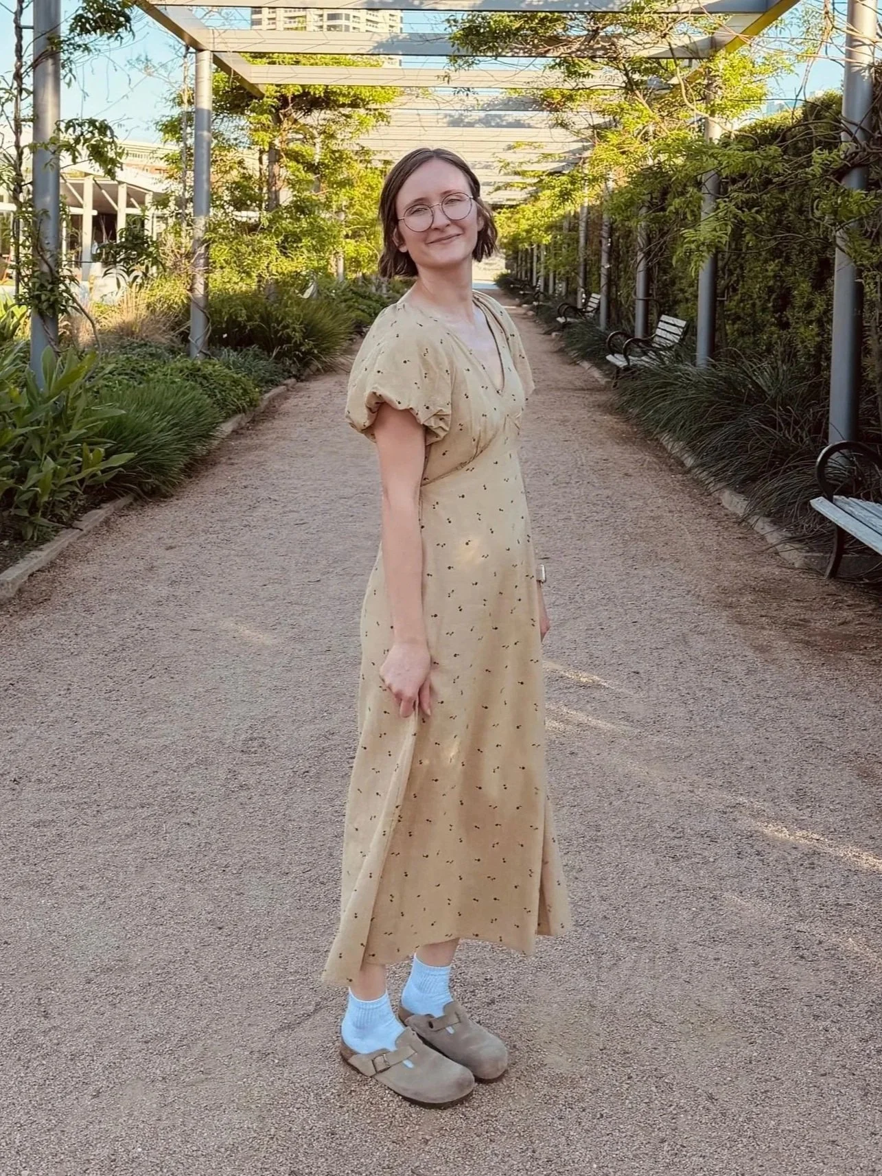 A young woman with short brown hair, glasses, and a light beige dress with small dark spots, standing on a gravel path in a green outdoor park. She is smiling and wearing beige Crocs with white socks. The background features plants, benches, and a metal pergola framework, with sunlight filtering through the trees.