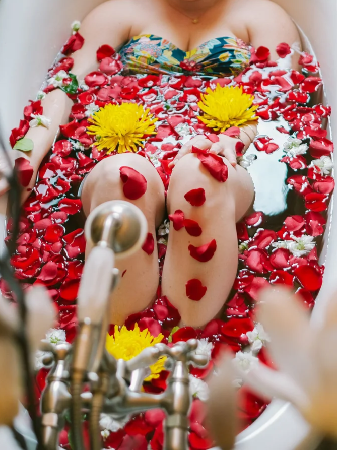 A person soaking in a bathtub filled with red rose petals and yellow flowers, wearing a colorful swimsuit, with their knees up and feet visible, surrounded by floral petals.