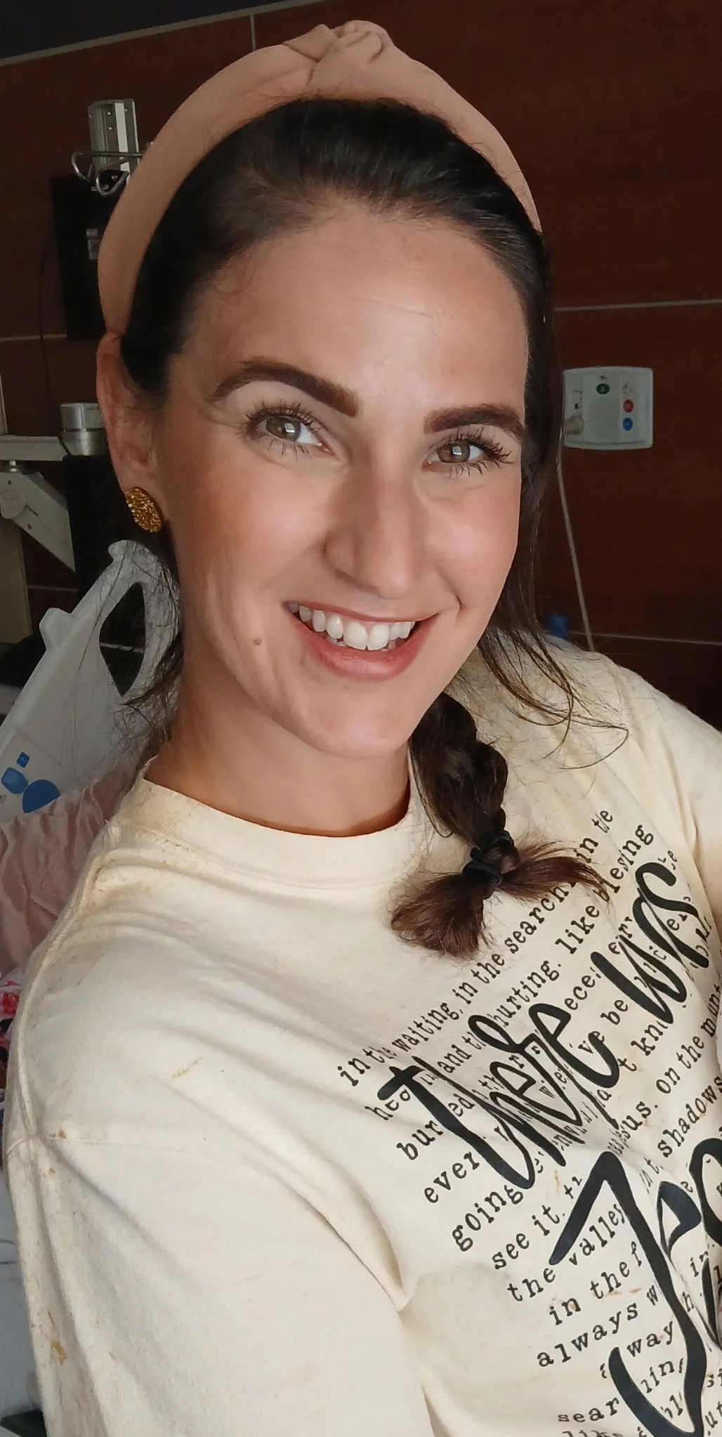 A smiling woman with dark hair in a braid, wearing a beige headband and gold earrings, taking a selfie in a medical or hospital room.