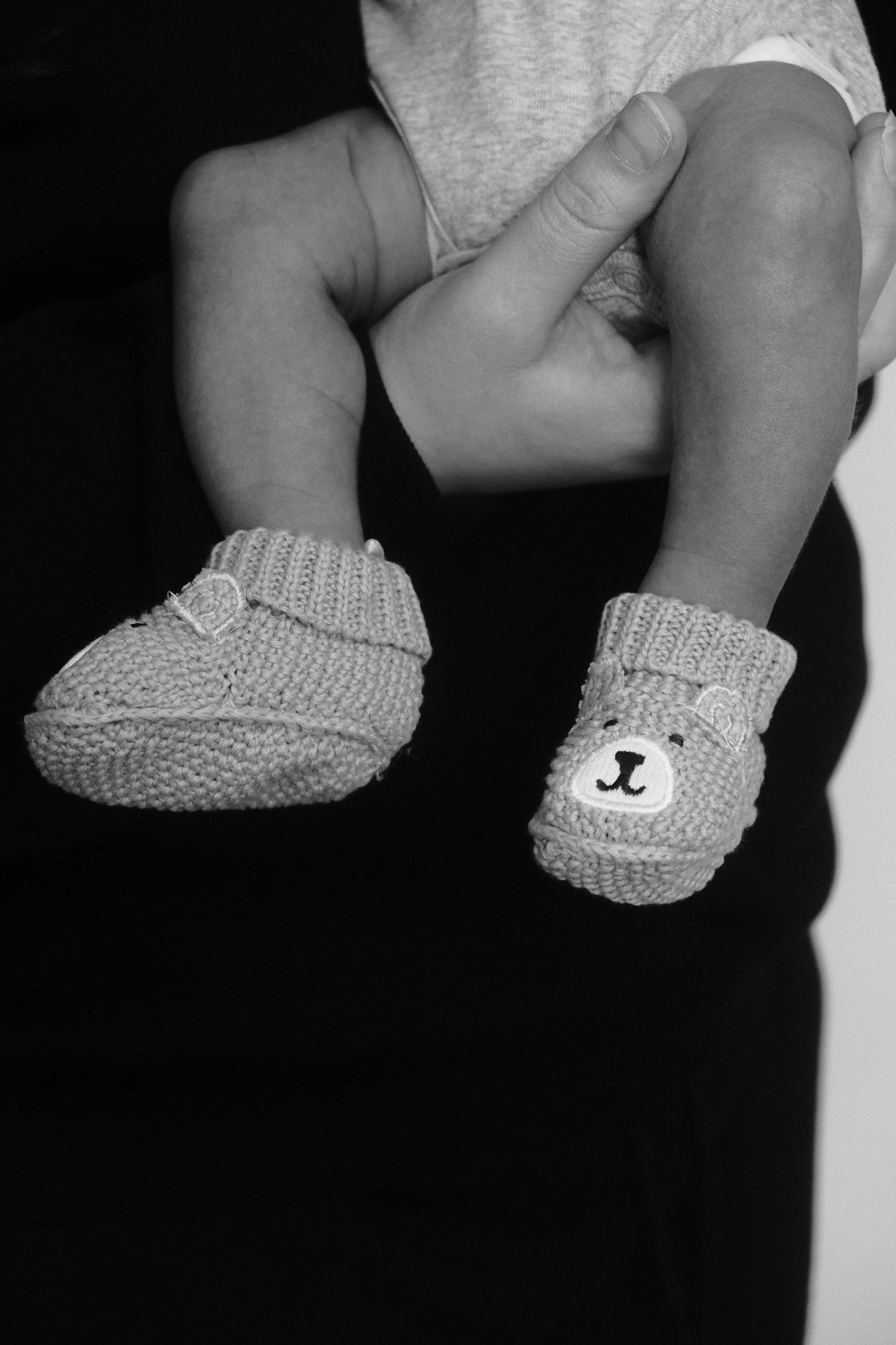 Close-up of a person's hands holding a small child, wearing knitted sleeves with bear faces on the cuffs, in black and white.