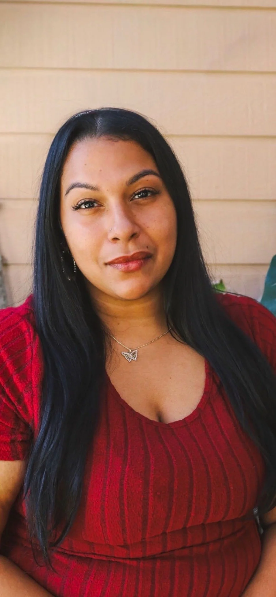 A woman with long black hair wearing a red top and butterfly necklace, sitting outdoors with a wooden wall in the background.