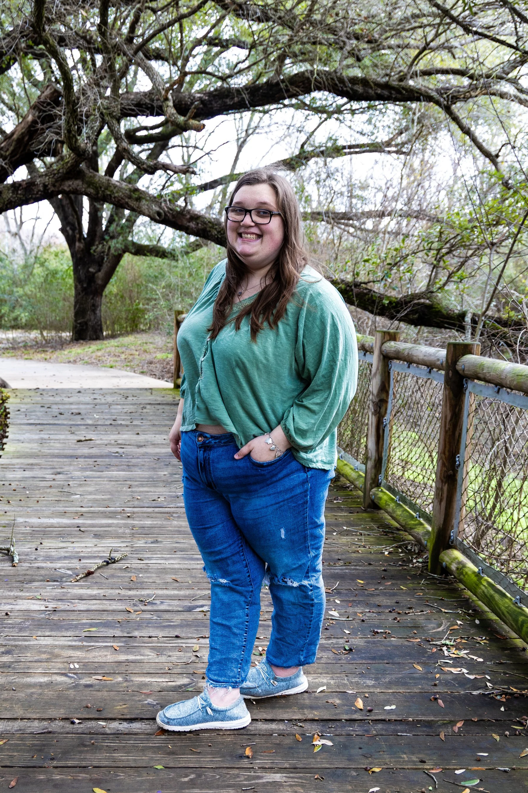 A young woman with long brown hair, glasses, and a bright smile standing on a wooden bridge outdoors, wearing a green cropped sweatshirt, ripped jeans, and sneakers, with trees and a cloudy sky in the background.