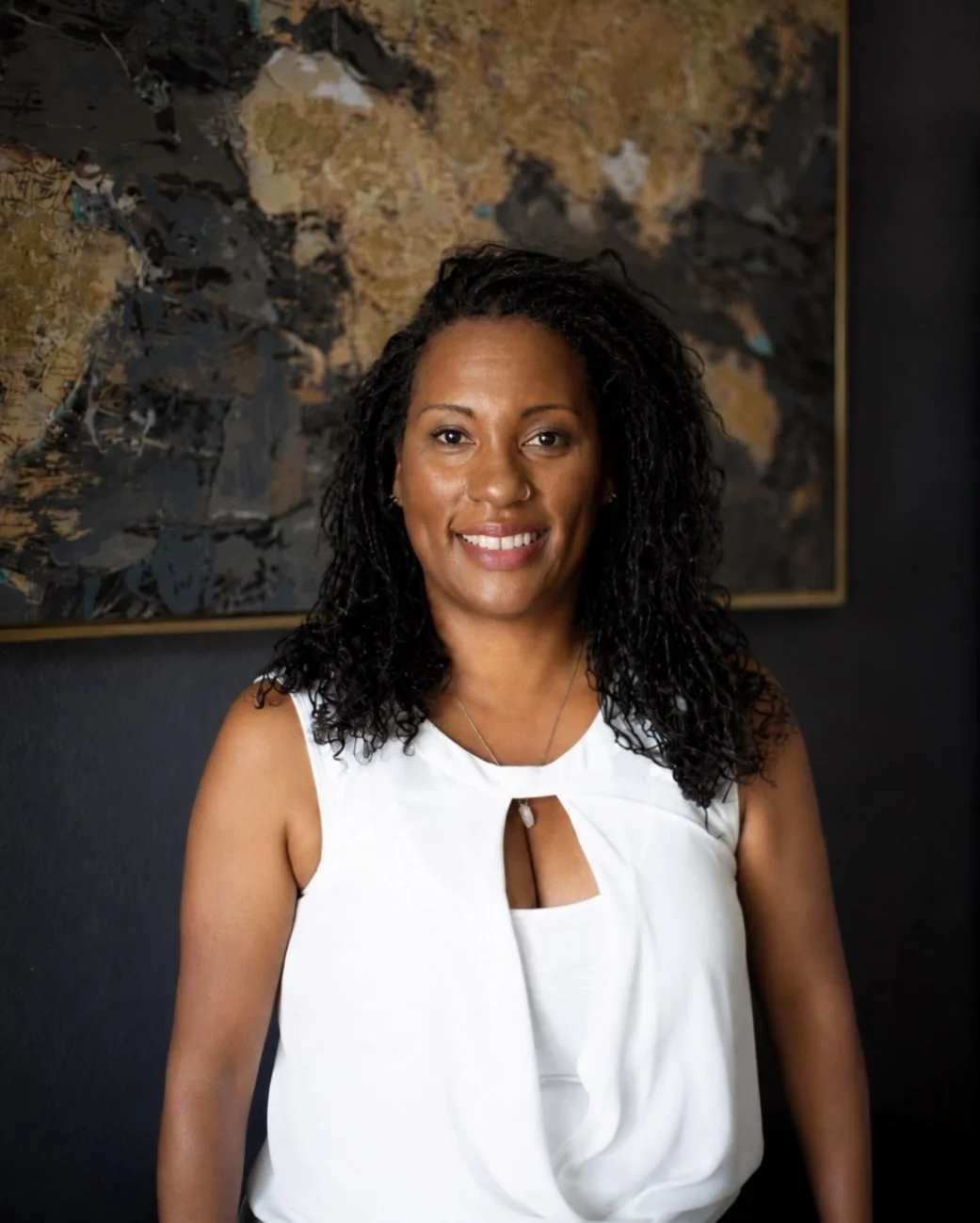 A smiling woman with curly black hair wearing a white sleeveless top with a keyhole cutout, standing in front of an abstract artwork on a dark wall.