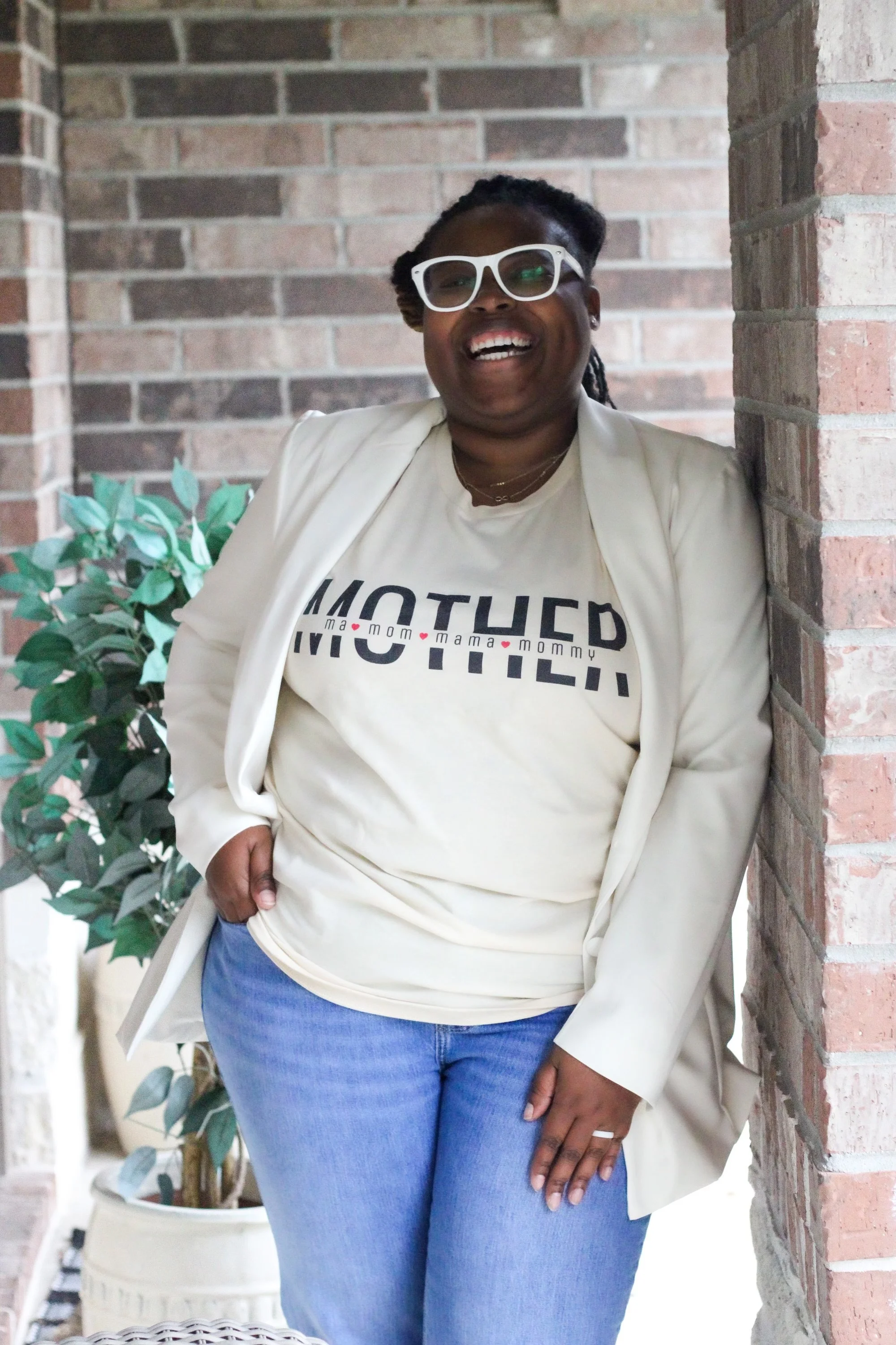 A woman with dark skin, wearing white glasses, a beige blazer, a graphic T-shirt, and blue jeans, smiling and standing against a brick wall near a potted plant.