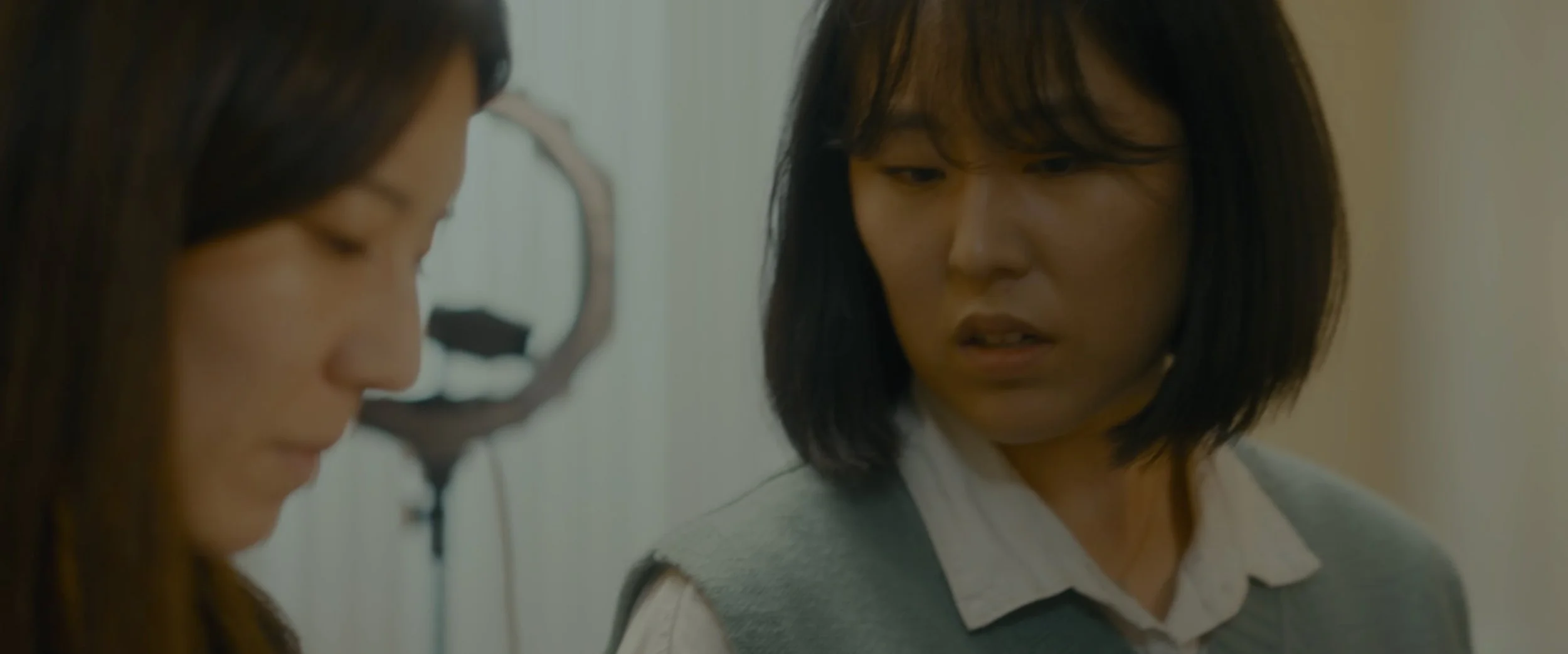 Two women with short, dark hair appear to be having a conversation; one of them is looking down, and the other is looking at her with a serious expression.