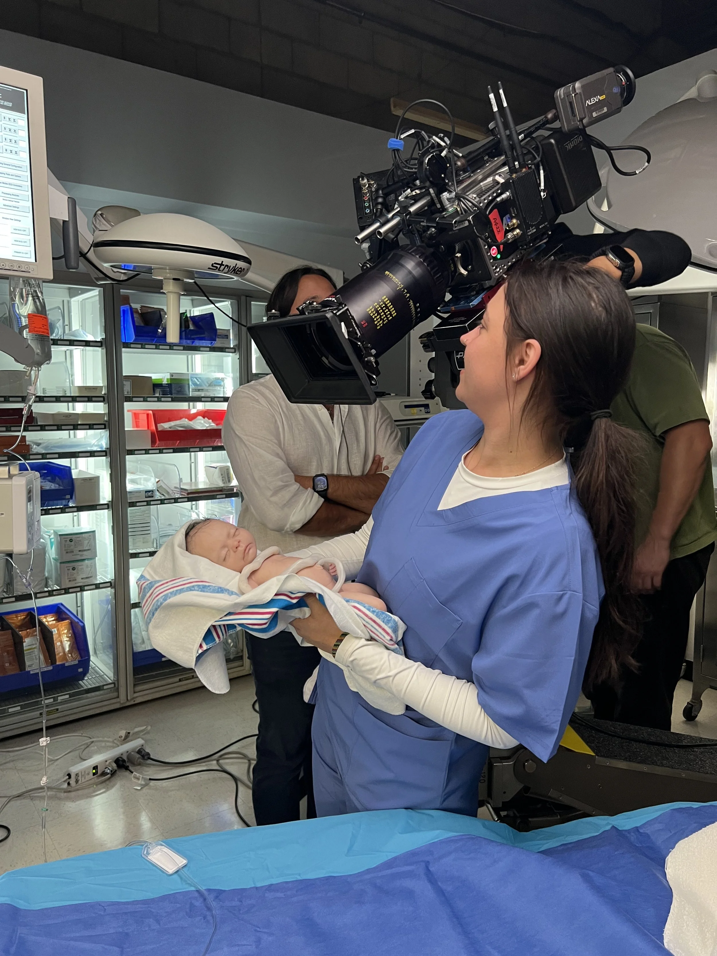 A female healthcare worker in scrubs holding a newborn baby in an hospital or medical setting while being filmed by a camera crew.