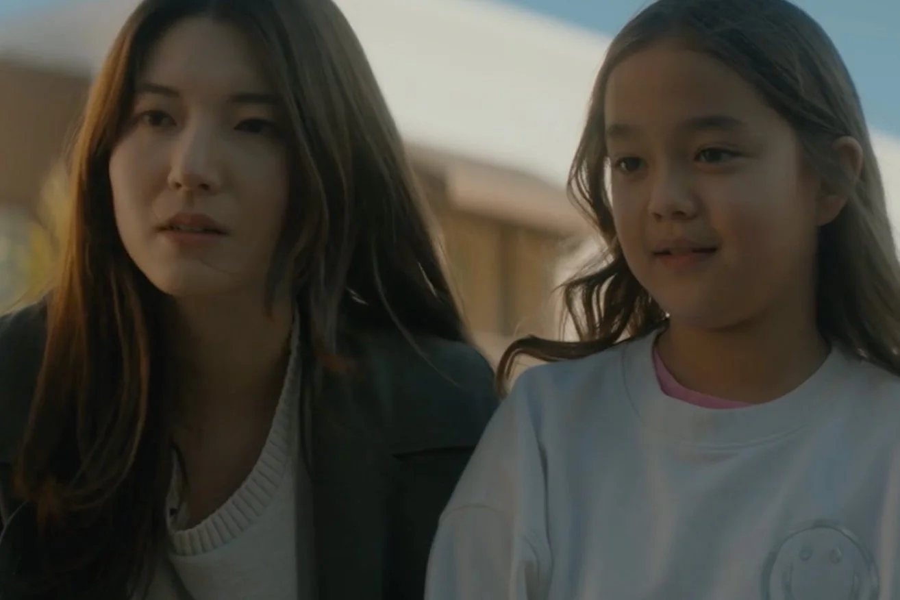 A woman and a young girl are outdoors, having a conversation and looking at something. The woman has long dark hair and is wearing a dark jacket, while the girl has long light brown hair and is wearing a white shirt with a small graphic.