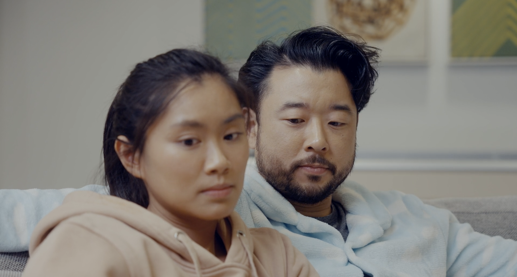 A man and woman sitting on a couch looking down with serious expressions.