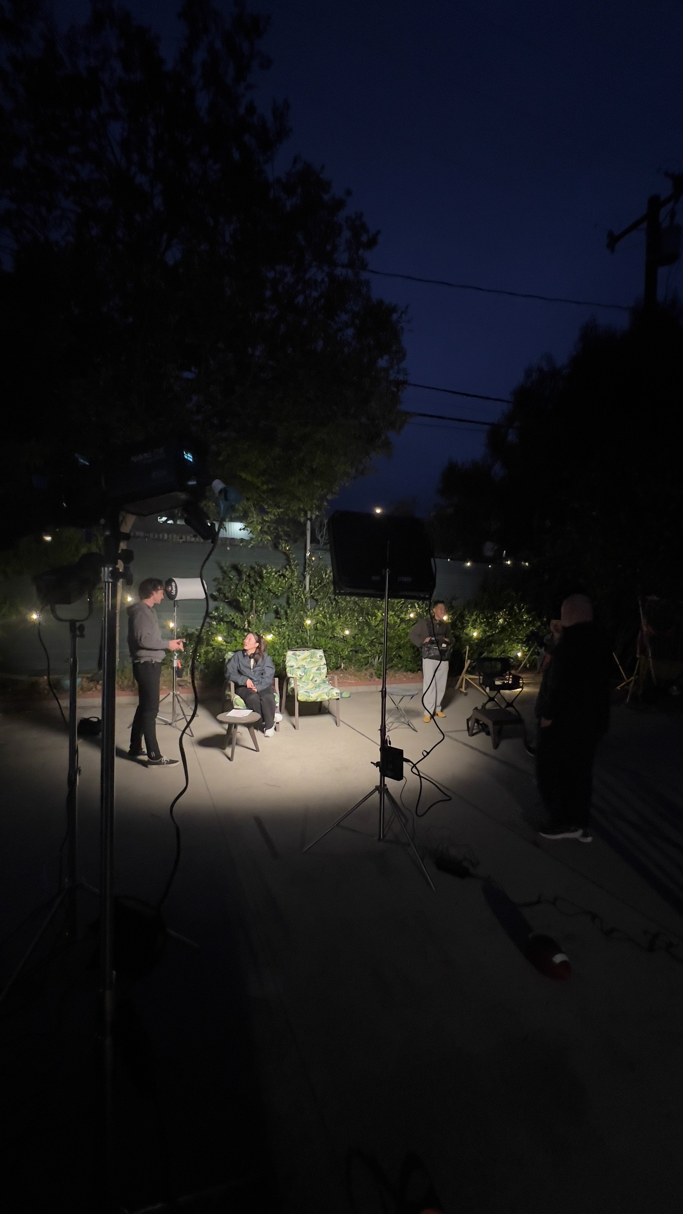 Nighttime outdoor film or video production set with crew members and lighting equipment, surrounded by trees and a dark sky.