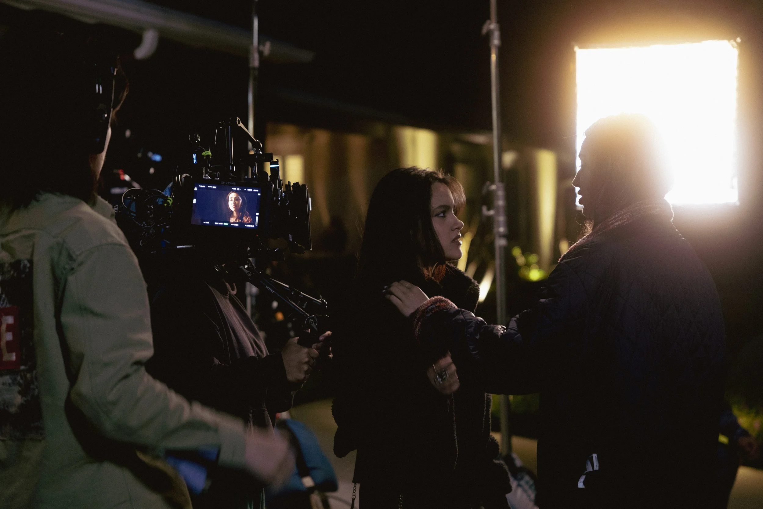 A behind-the-scenes photo of a film or photography crew filming an outdoor scene at night, with a woman facing a bright light source, and crew members operating equipment and camera.