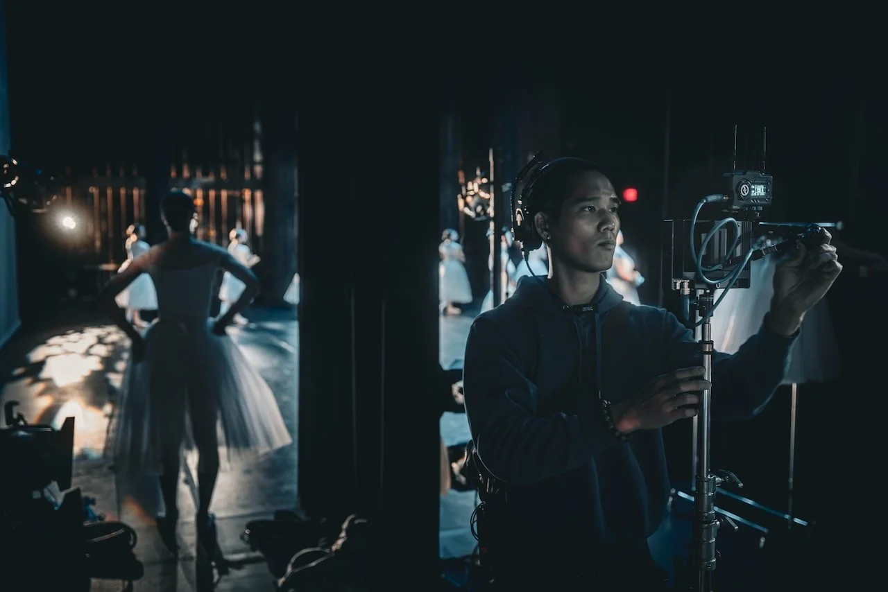 Behind-the-scenes photo of a ballet dancer on stage with other dancers in background, and a crew member operating filming equipment in the foreground.