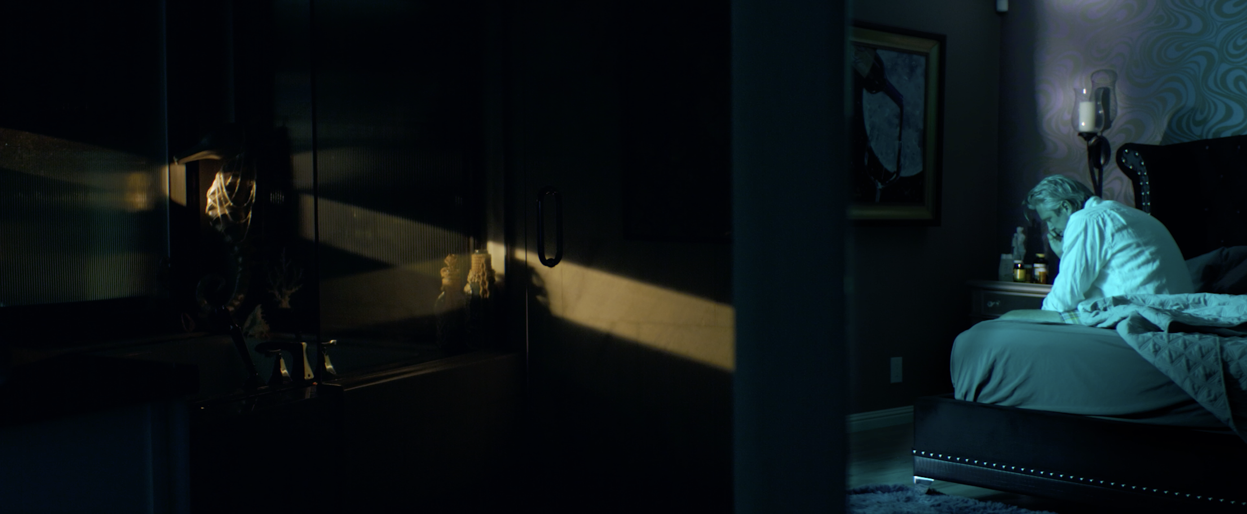 A person sitting on a bed in a dimly lit bedroom, illuminated by soft nightlight and shadows.