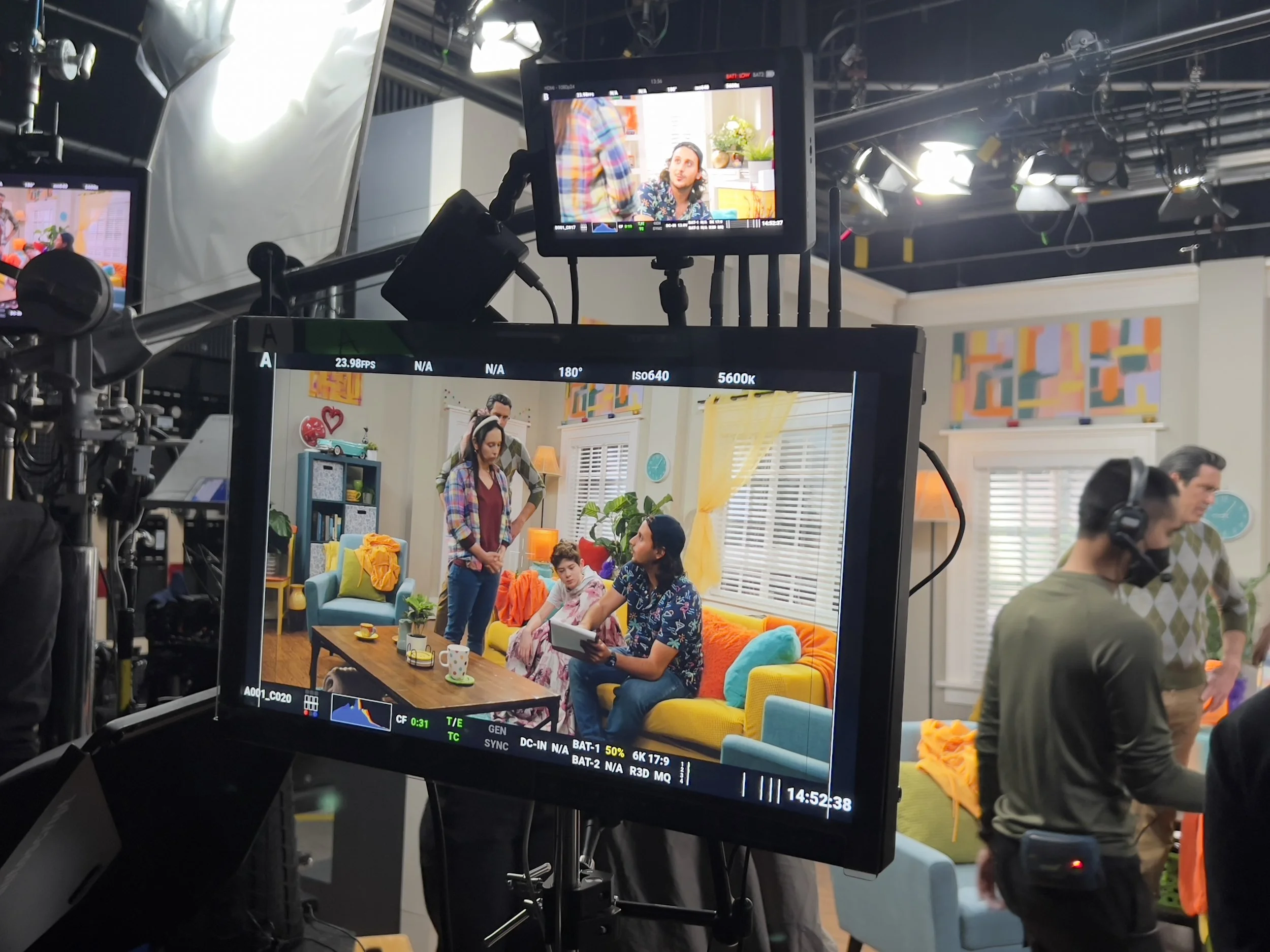 View of a television camera monitor capturing a colorful living room scene where people are sitting and standing, with a woman in a plaid shirt and a man with long hair wearing a flowered shirt, among vibrant furniture and decorations.