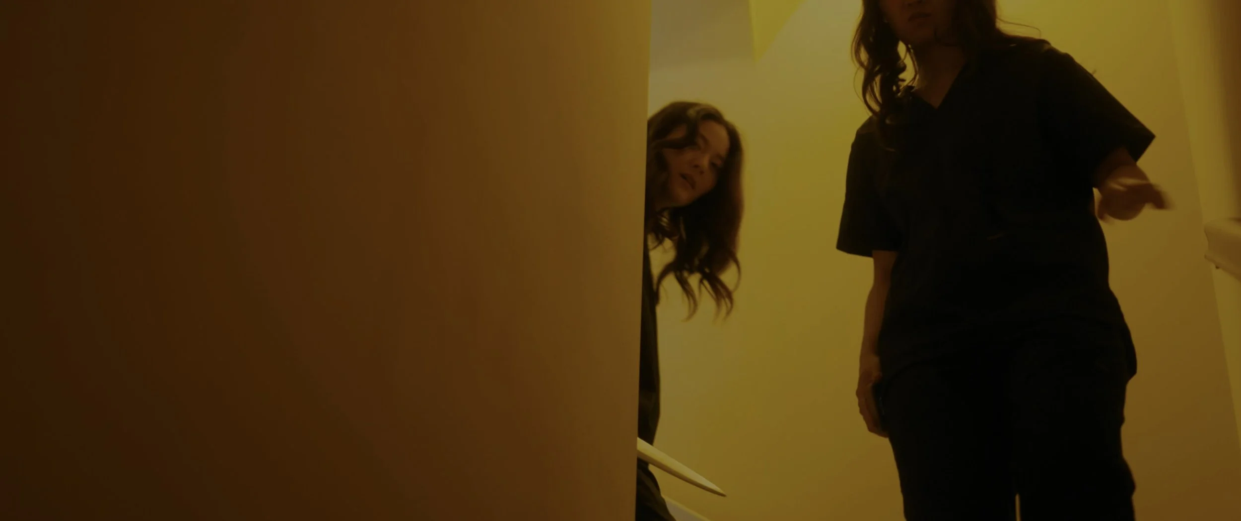 Two women in black clothing in a dimly lit yellow hallway, one woman leaning against a wall and the other standing with her hand extended.