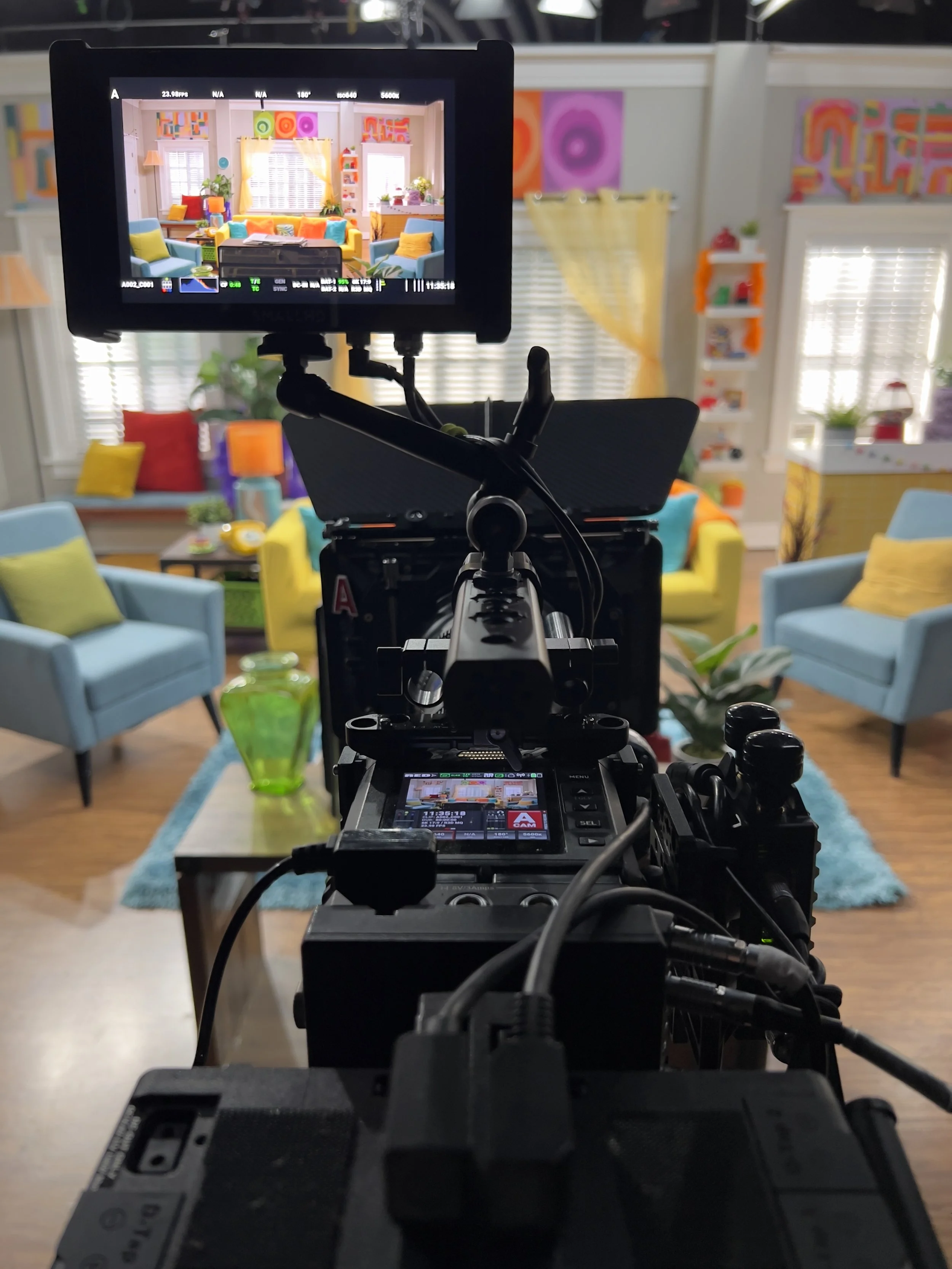 A professional video camera on a tripod recording a colorful, modern living room with blue and yellow armchairs, vibrant cushions, plants, and colorful abstract art on the walls.