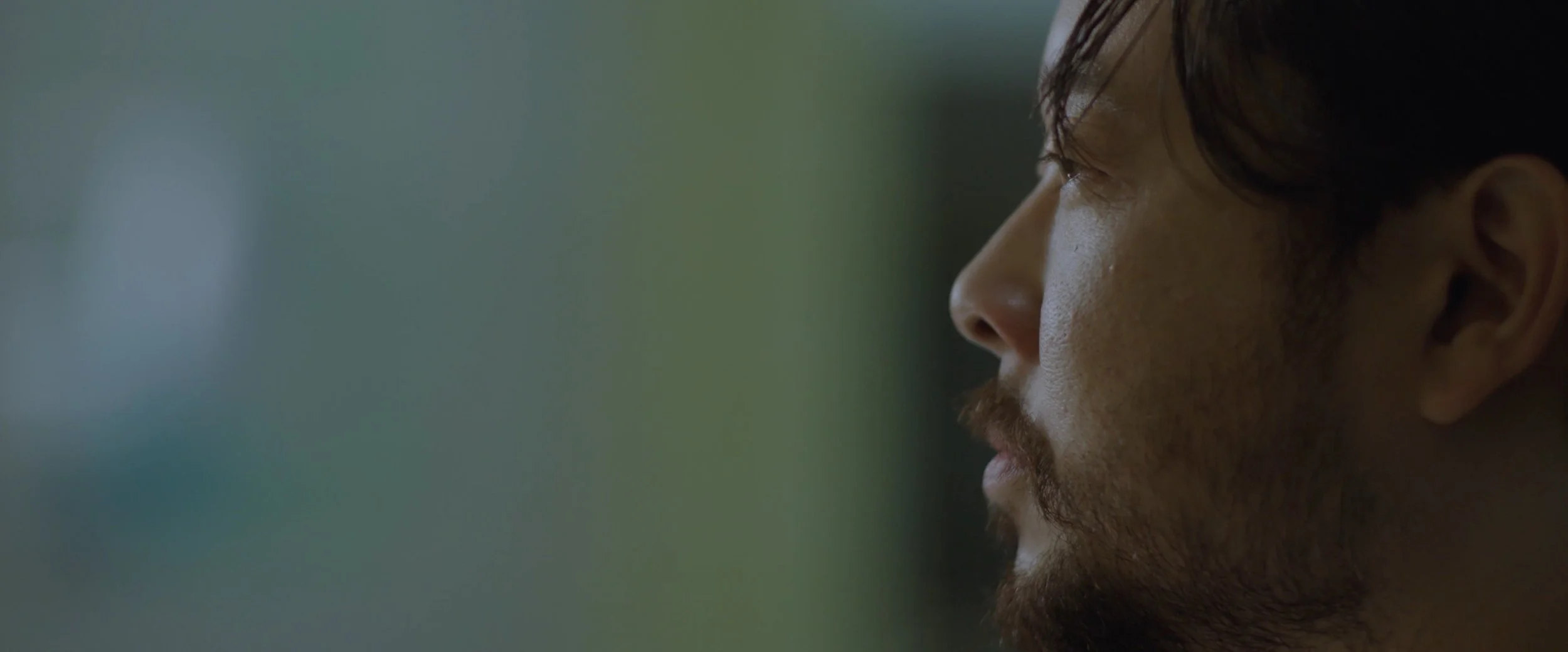 Close-up profile of a man with dark hair, beard, and mustache, looking to the right in a dimly lit setting.