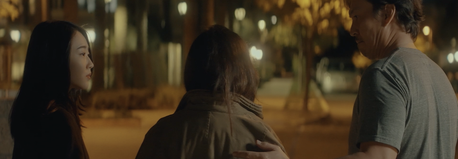 Three people standing outside at night having a conversation, with trees and lights in the background.