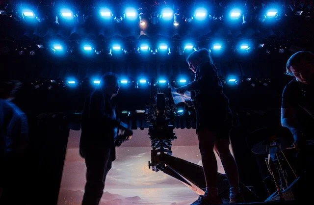 Behind the scenes of a production with crew members and equipment in a dark studio with blue lights overhead.