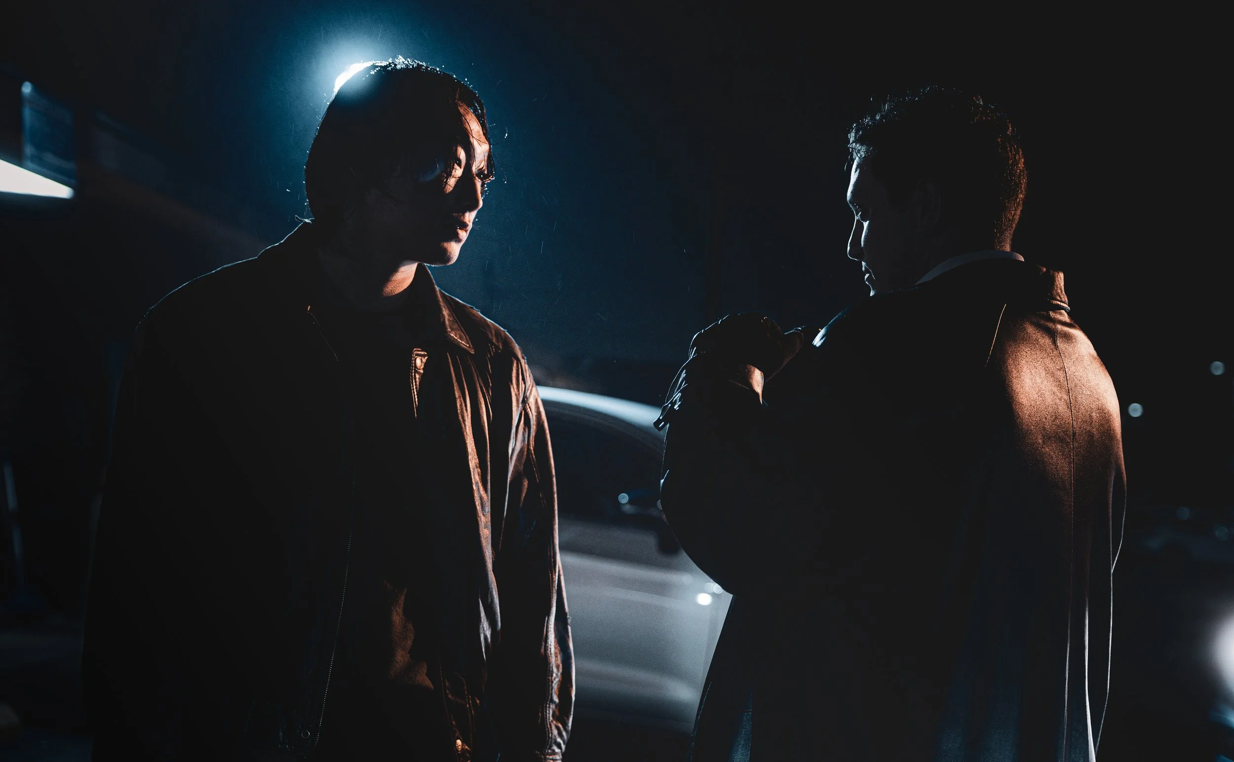 Two men in leather jackets face each other in a dark, rainy night, illuminated by a backlight.