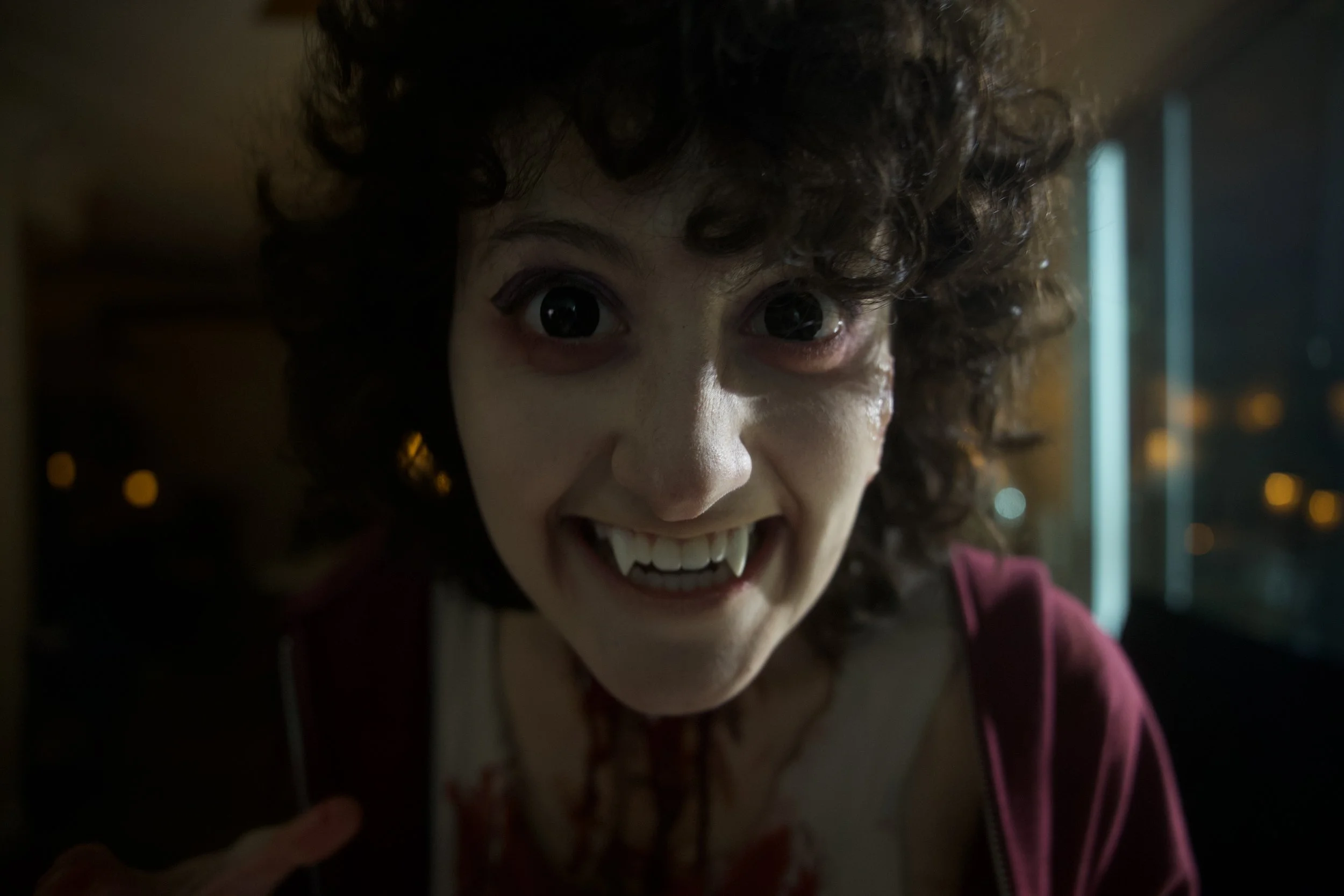 Close-up of a person with curly hair, wide eyes, sharp teeth, and a creepy smile, wearing a blood-stained shirt, inside a dimly lit room at night.