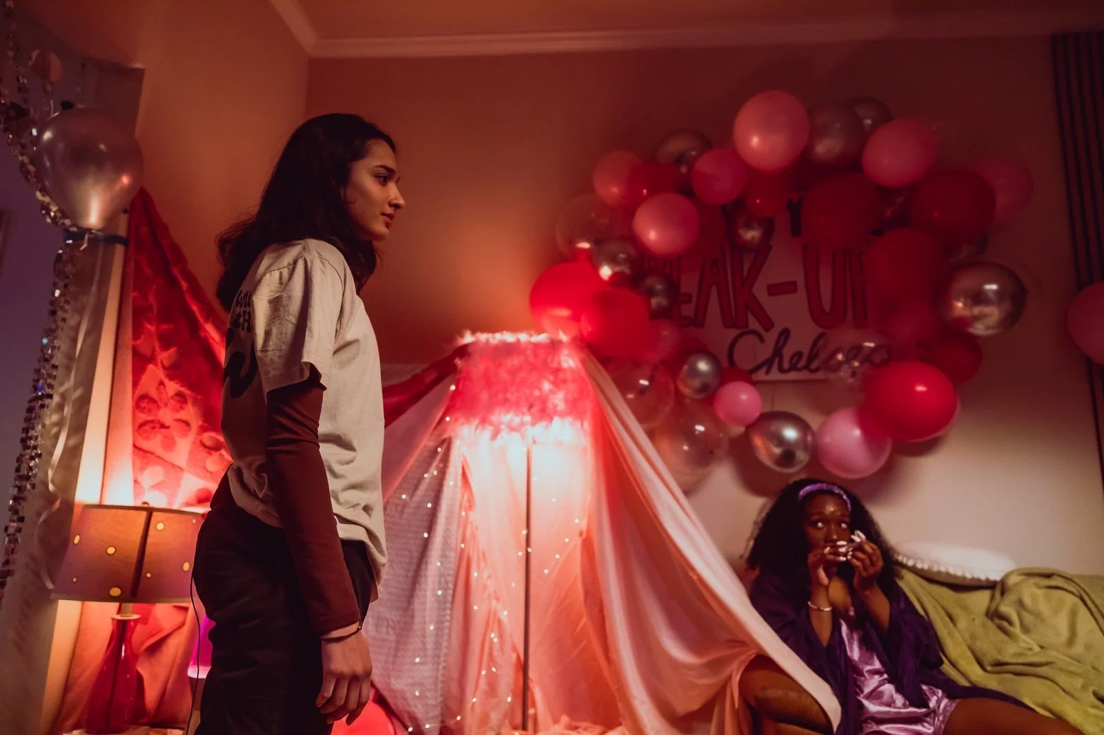 A woman stands in a decorated room with pink and silver balloons, a pink canopy, and a pink-lit lamp; another woman sits on a couch.