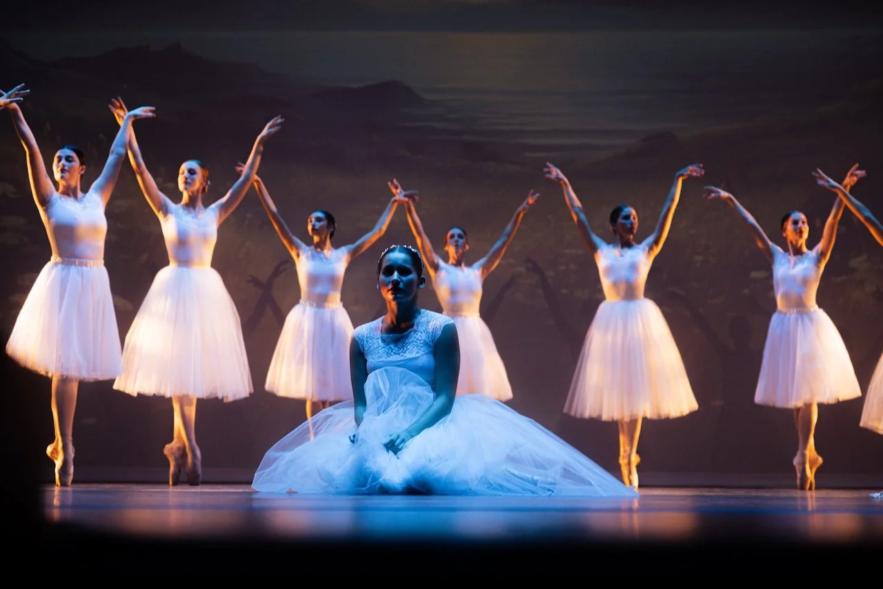 Ballet dancers on stage, with one dancer kneeling in front wearing a white tutu and the others standing behind with their arms raised