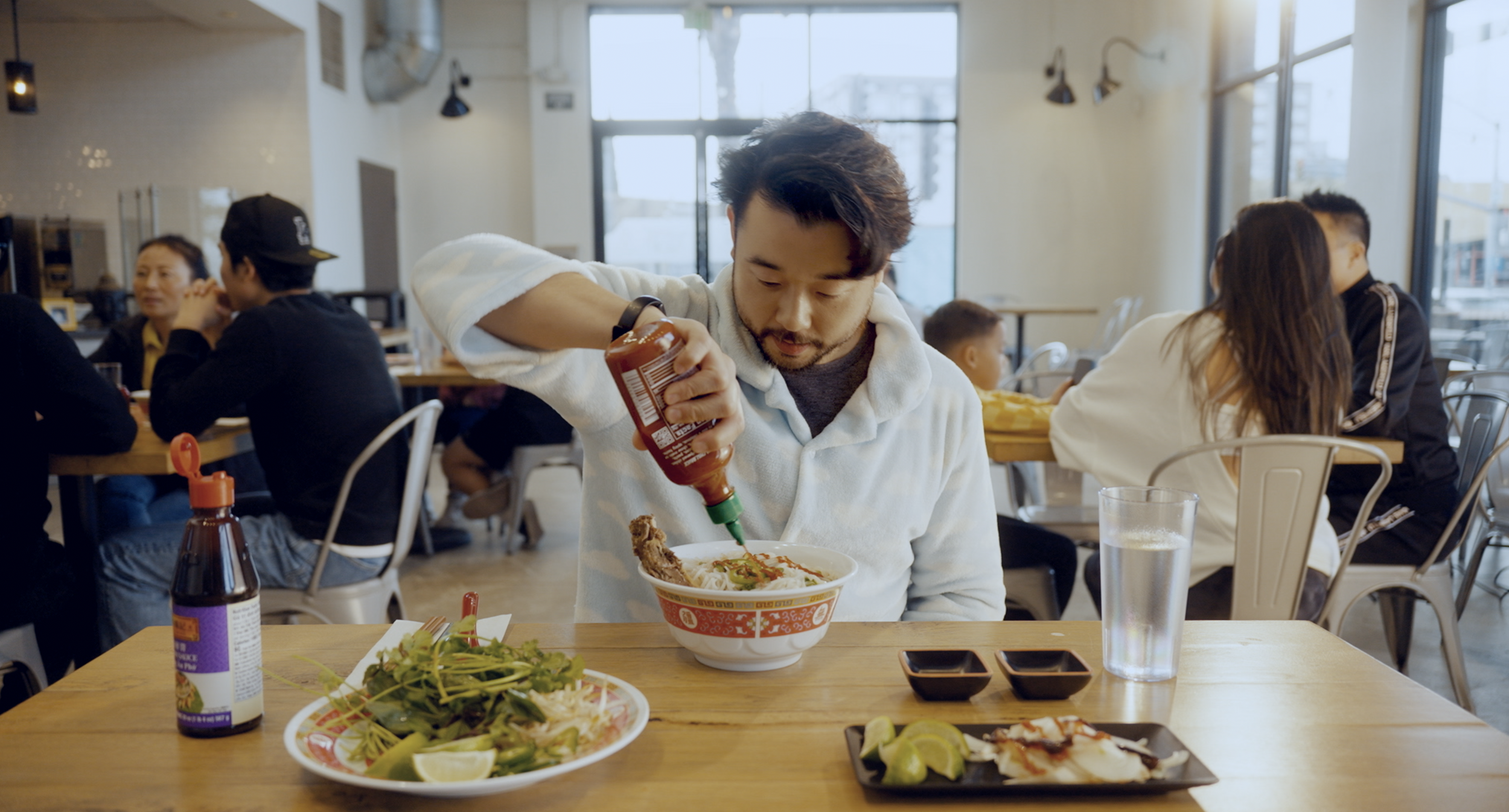 A man is pouring hot sauce onto a bowl of ramen at a restaurant. On the table are salads, lime slices, and dipping sauces.
