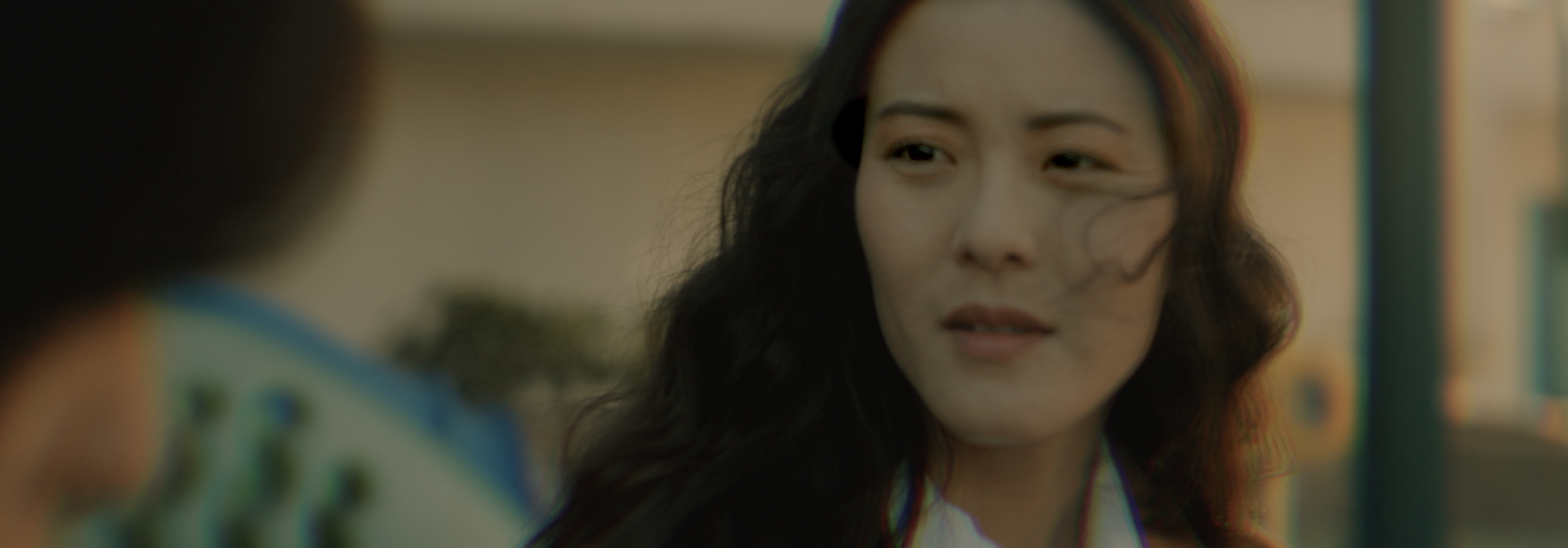 Close-up of a woman's face with long dark hair, looking slightly to the side, outdoors with a blurred background.