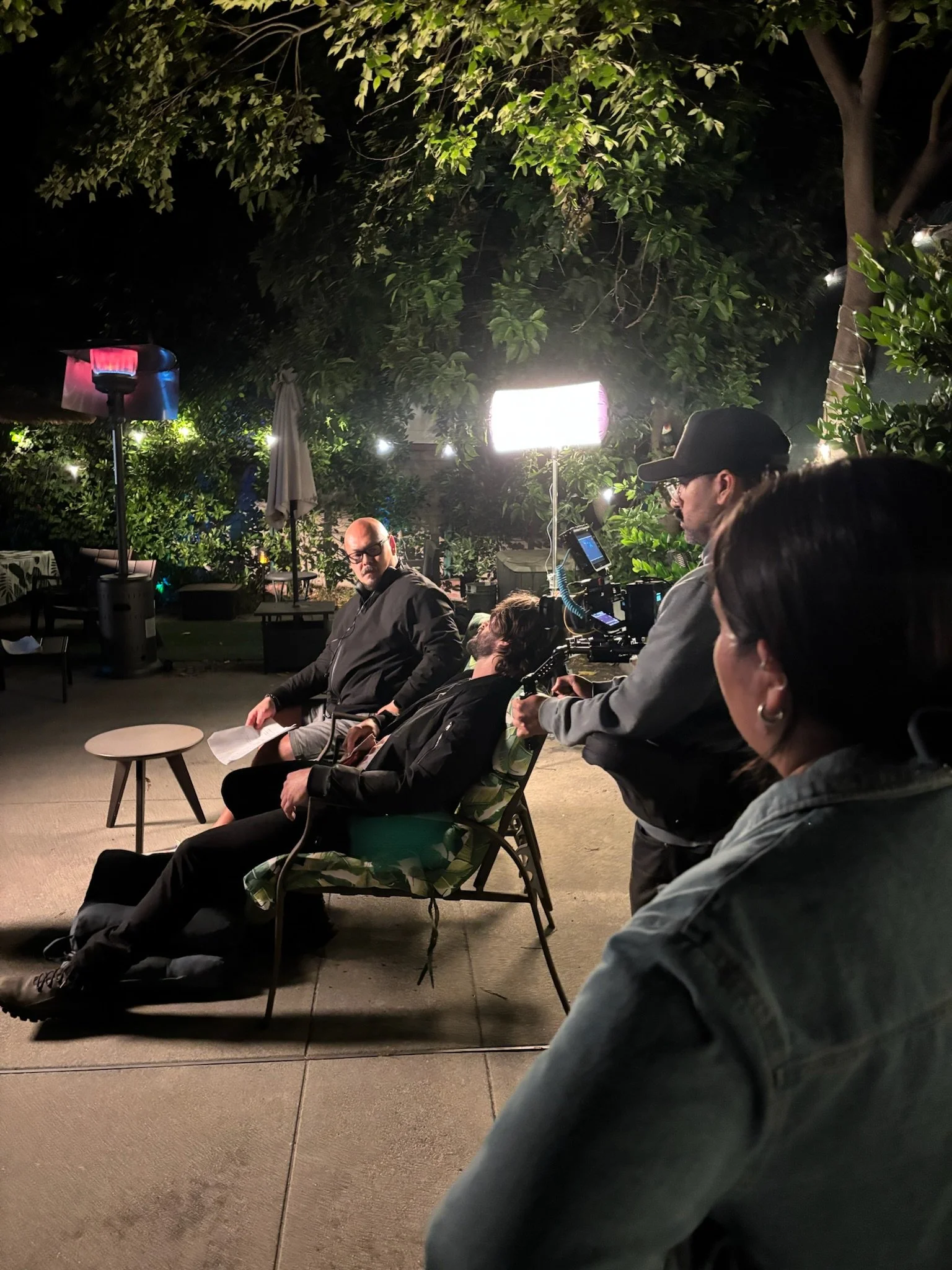 People sitting outdoors at night, with lighting equipment and greenery around.