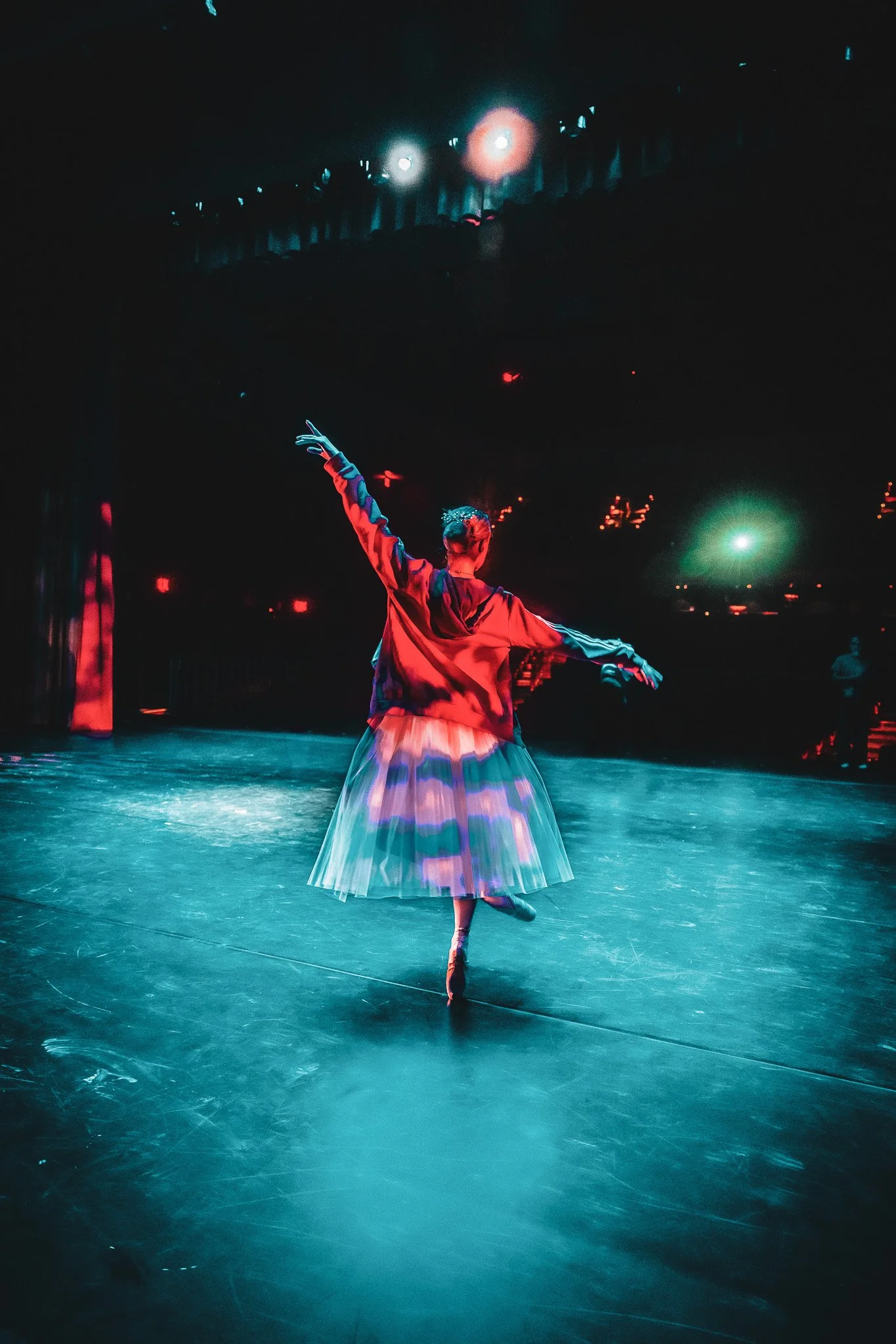 Ballet dancer performing on a stage under stage lights, wearing a colorful costume with a pleated skirt and a loose top.