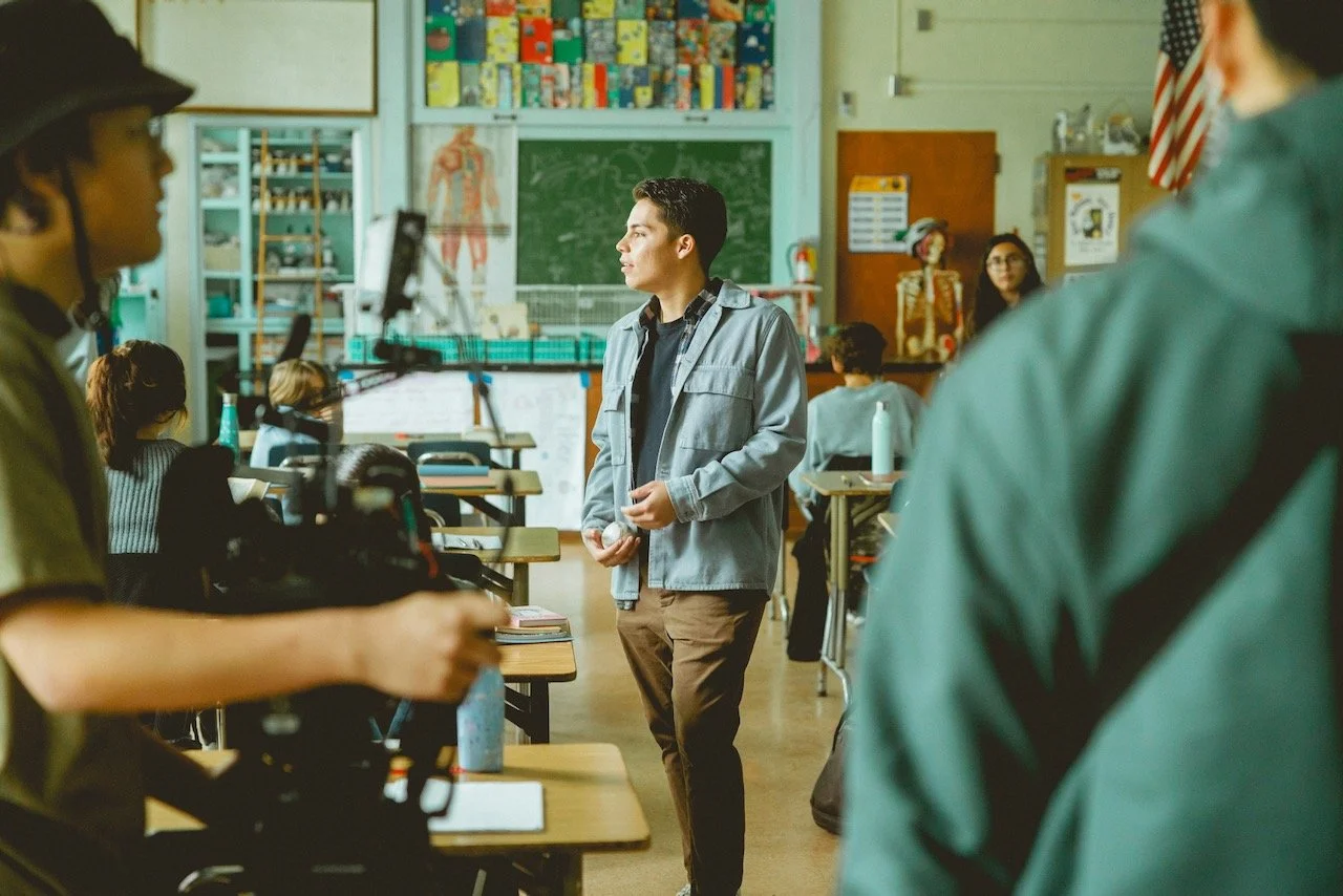 A classroom with students and a man standing at the front of the room. The classroom has educational posters, a chalkboard, and various supplies.