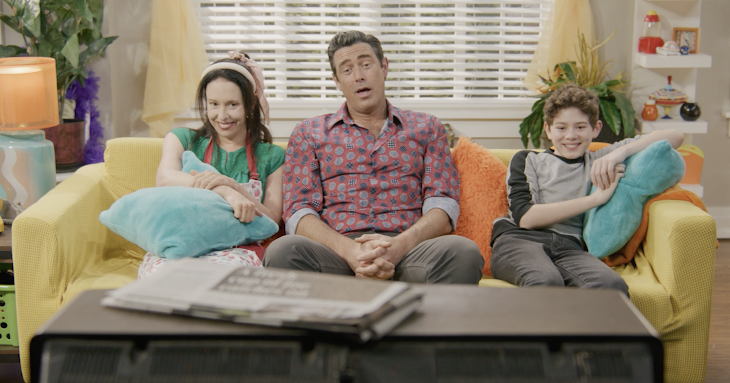 Three people sitting on a yellow couch watching TV in a colorful living room. An adult man in the middle, a woman on the left with dark hair and a woman on the right with short curly hair, both smiling.
