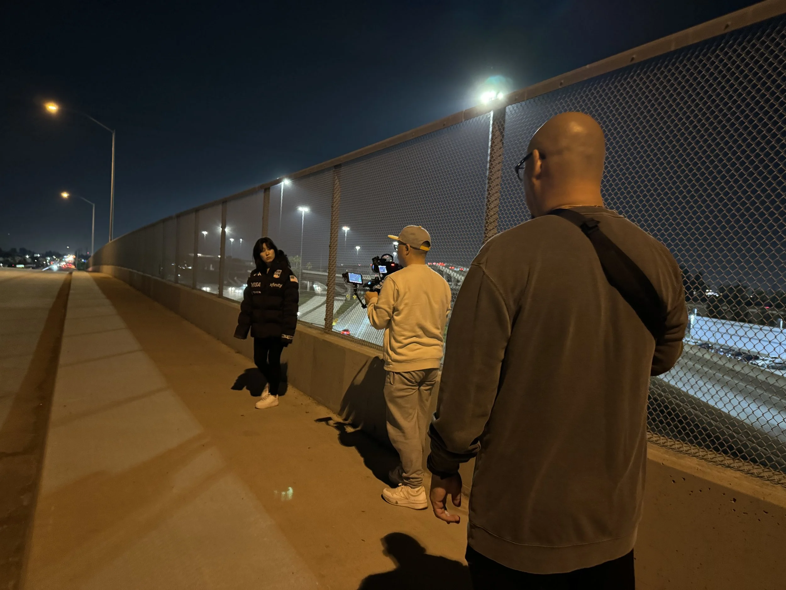 A group of four people standing on a sidewalk beside a chain-link fence at night. One person is facing a camera, and the others are turned away or partially visible. City lights are visible in the background.