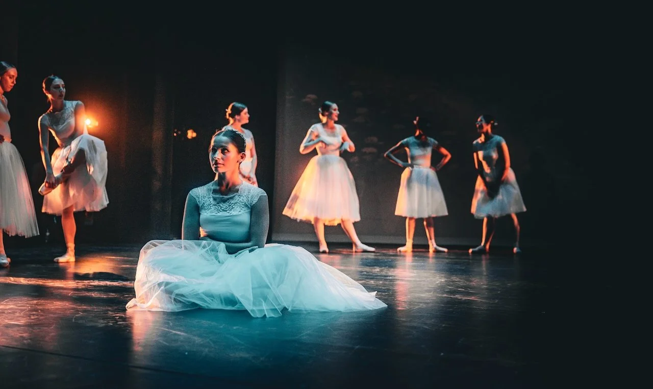Ballet dancers on stage, with one seated in the foreground and five standing in the background, performing in a dimly lit theater.