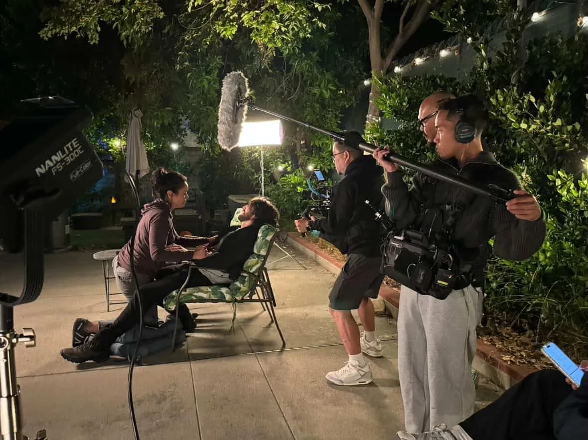 Filming scene of two women talking in a backyard at night, filming equipment including a camera, boom mic, and crew members present.