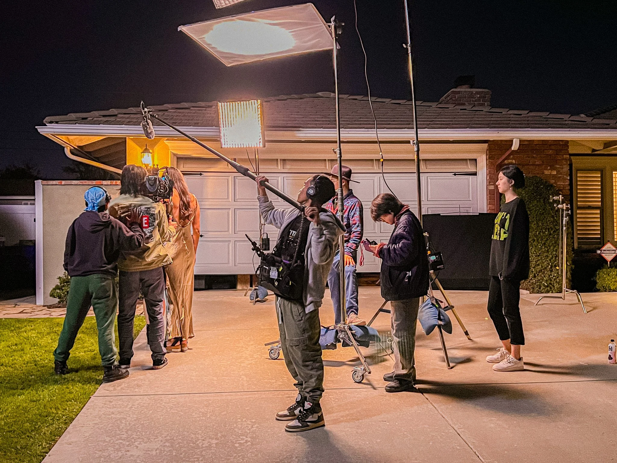 Film crew filming a scene outside a house at night, with lighting equipment and crew members operating cameras and gear.