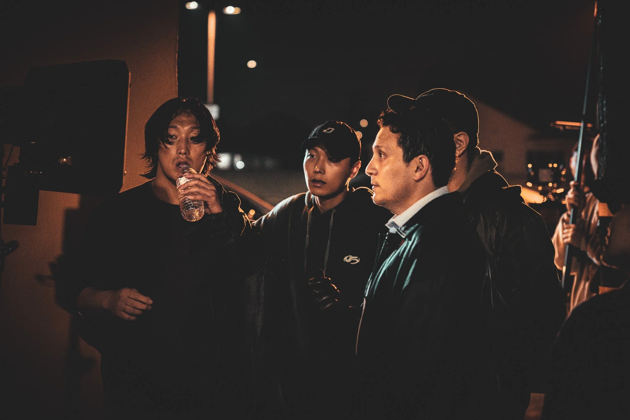 Group of young men on film set at night, with one drinking water and others listening intently.