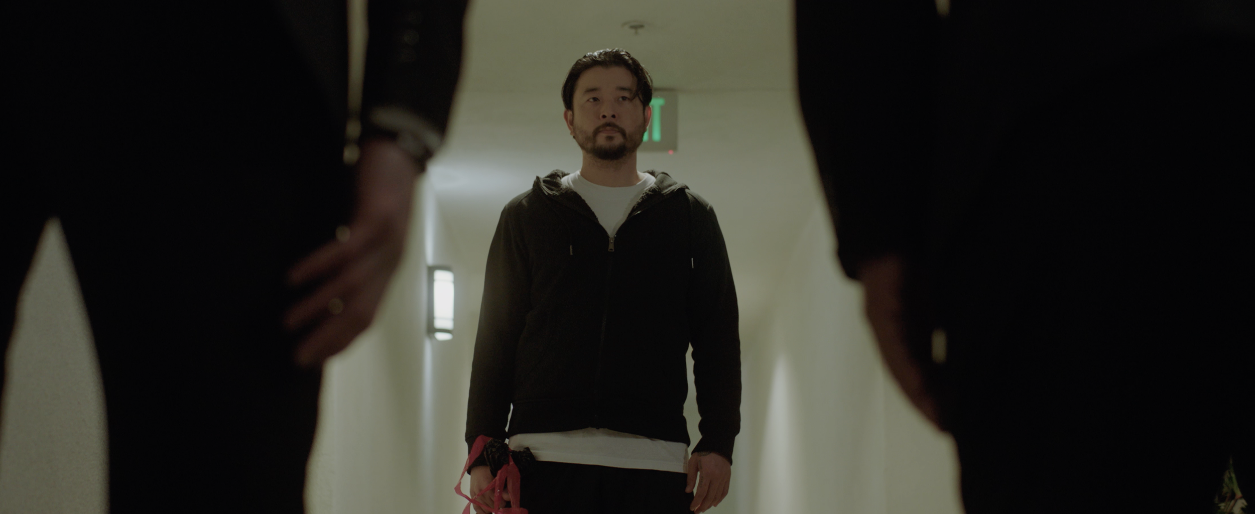 Man standing between two people whose legs are in the foreground, viewed from a low angle in a hallway.