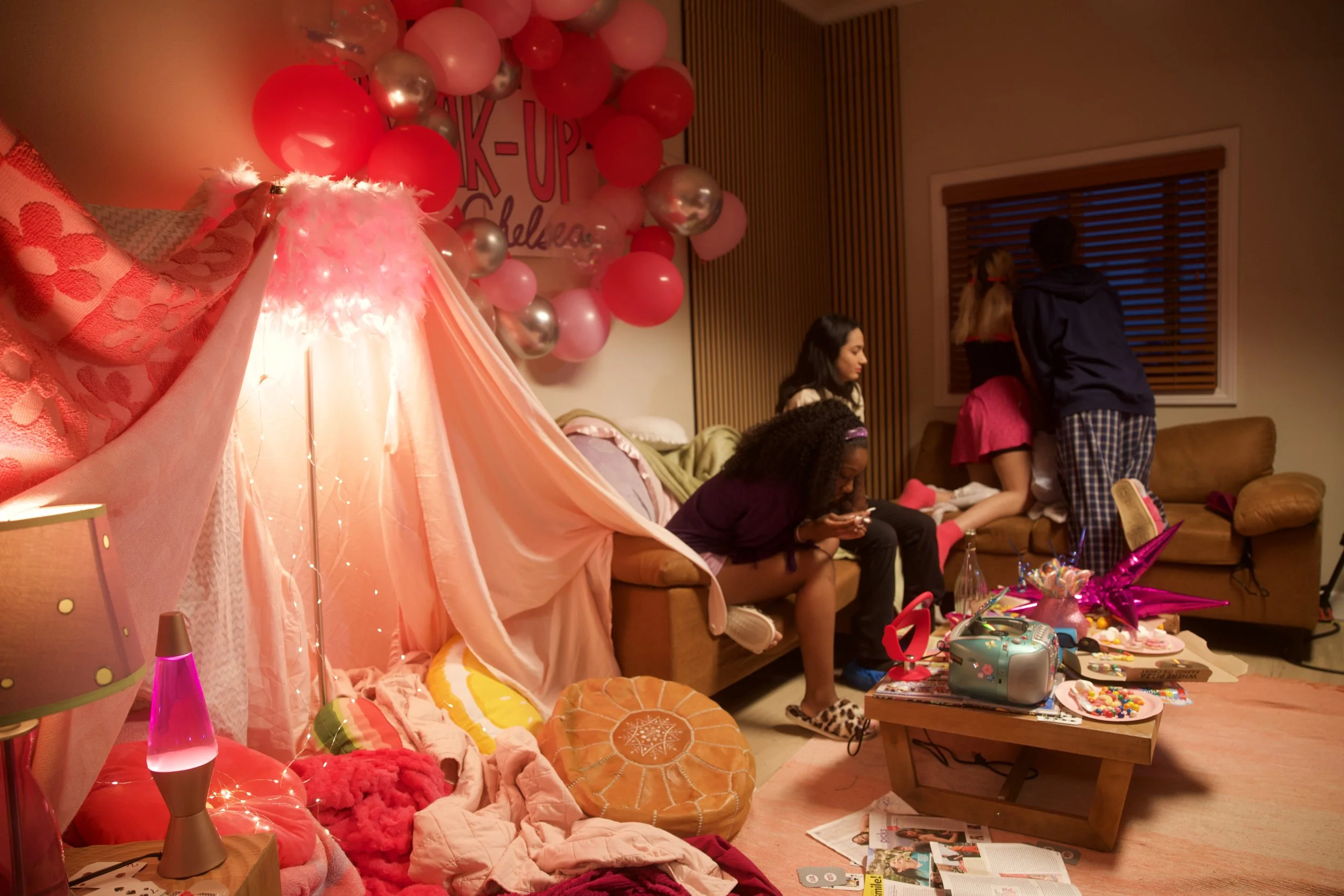 A cozy room decorated with pink and silver balloons, fairy lights, and pink fabric drapes. Four young women are in the room, one sitting on the couch engaged with her phone, one kneeling on the floor, and two standing near a window, engaging in conve