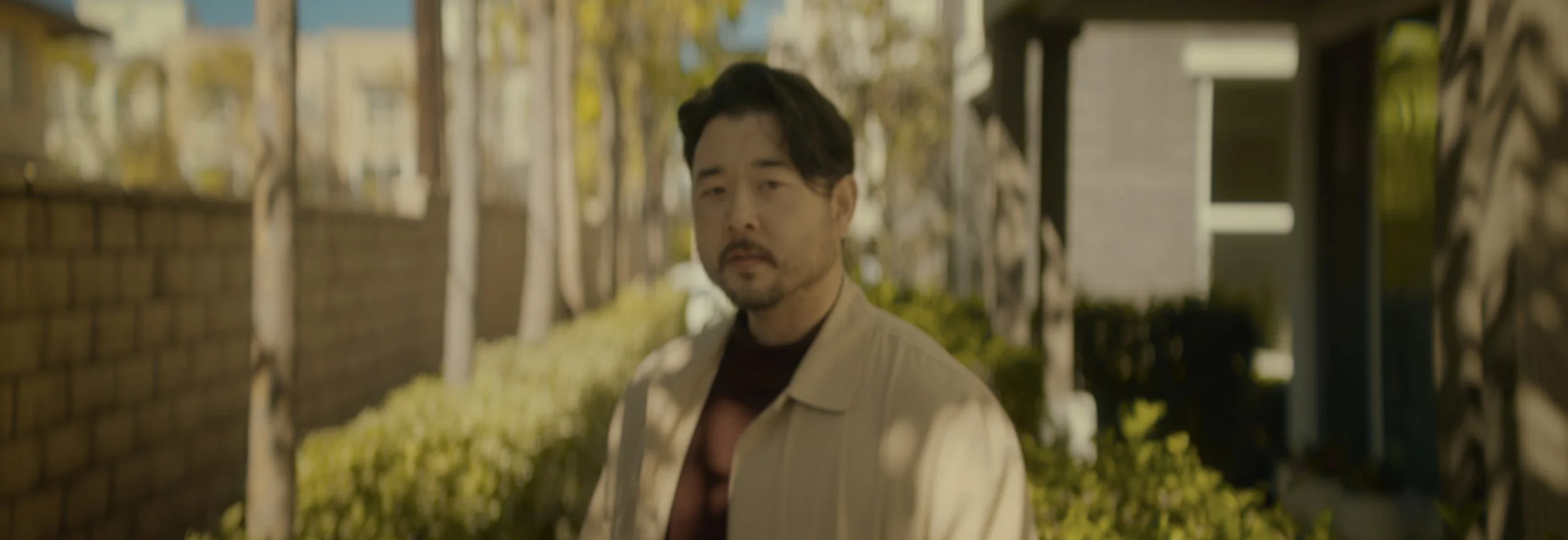 A man with black hair and facial hair wearing a beige jacket standing outdoors in a neighborhood with trees and houses in the background.