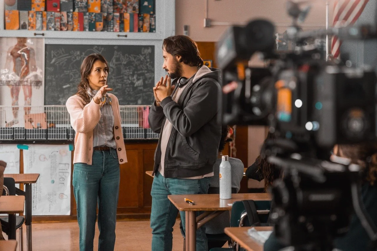 A woman and a man having a conversation in a classroom or science lab, with a camera filming them in the foreground.