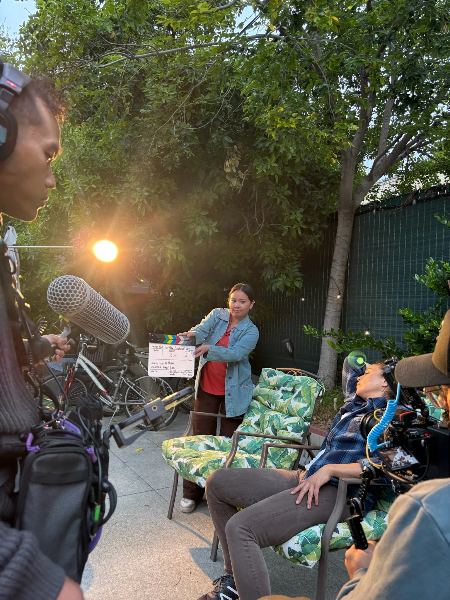 A woman in a denim jacket holds a film slate, surrounded by people with cameras and microphones, outdoors with green trees and a black fence in the background.