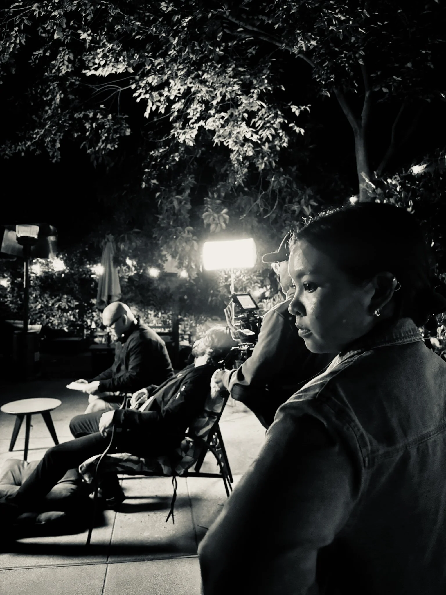 People sitting on chairs outdoors at night, some looking at their phones, with trees and lights in the background.